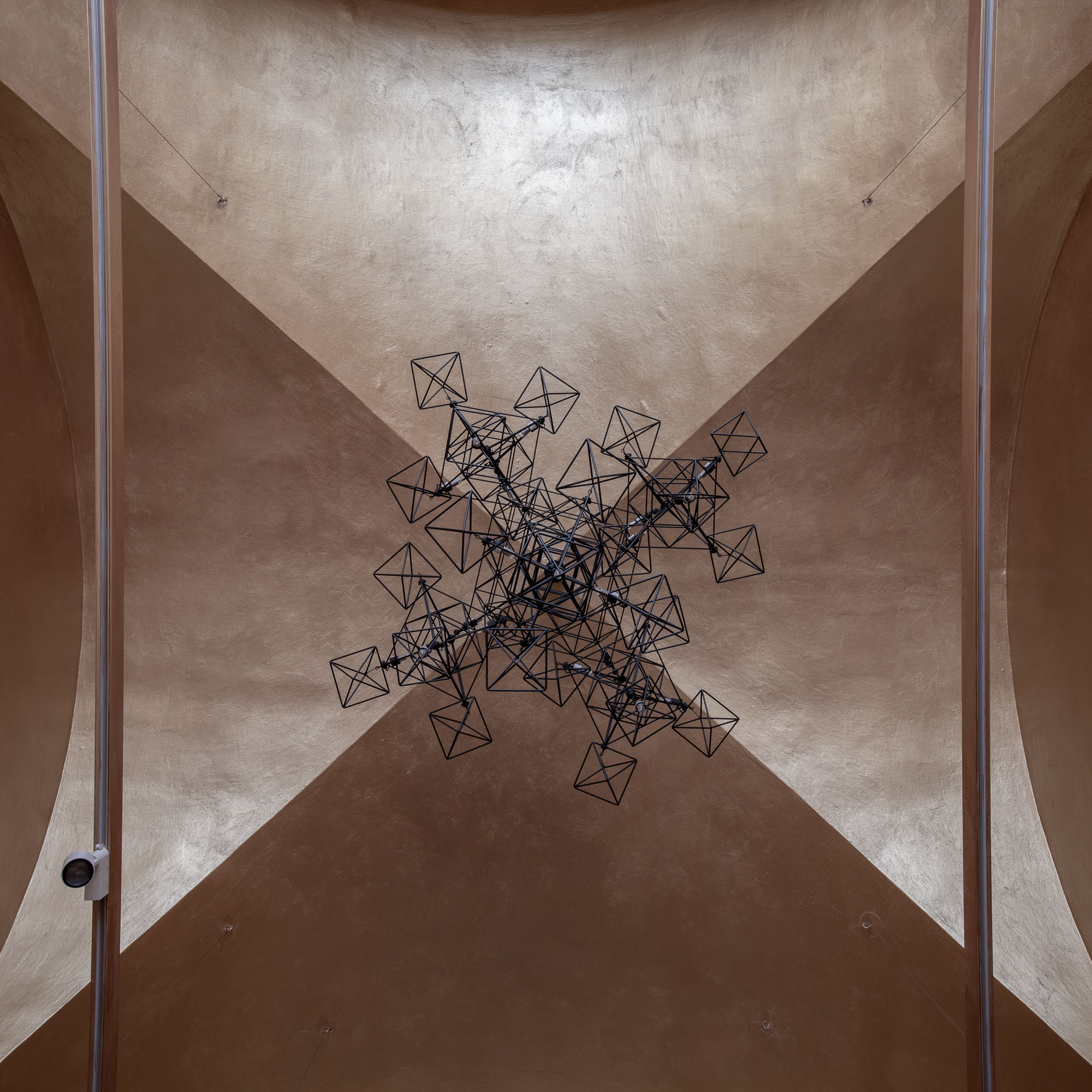 Pohled nahoru na zářivé geometrické drátěné svítidlo v recepci galerie ARCIMBOLDO. Shluky oktaedrických tvarů na zlaté klenbě. Praha.