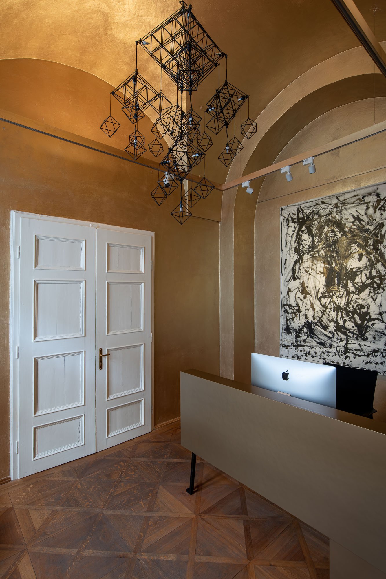 Drátěné geometrické svítidlo v recepci galerie ARCIMBOLDO fotografované zdola. Oktaedrické tvary na zlaté texturované omítkové klenbě.