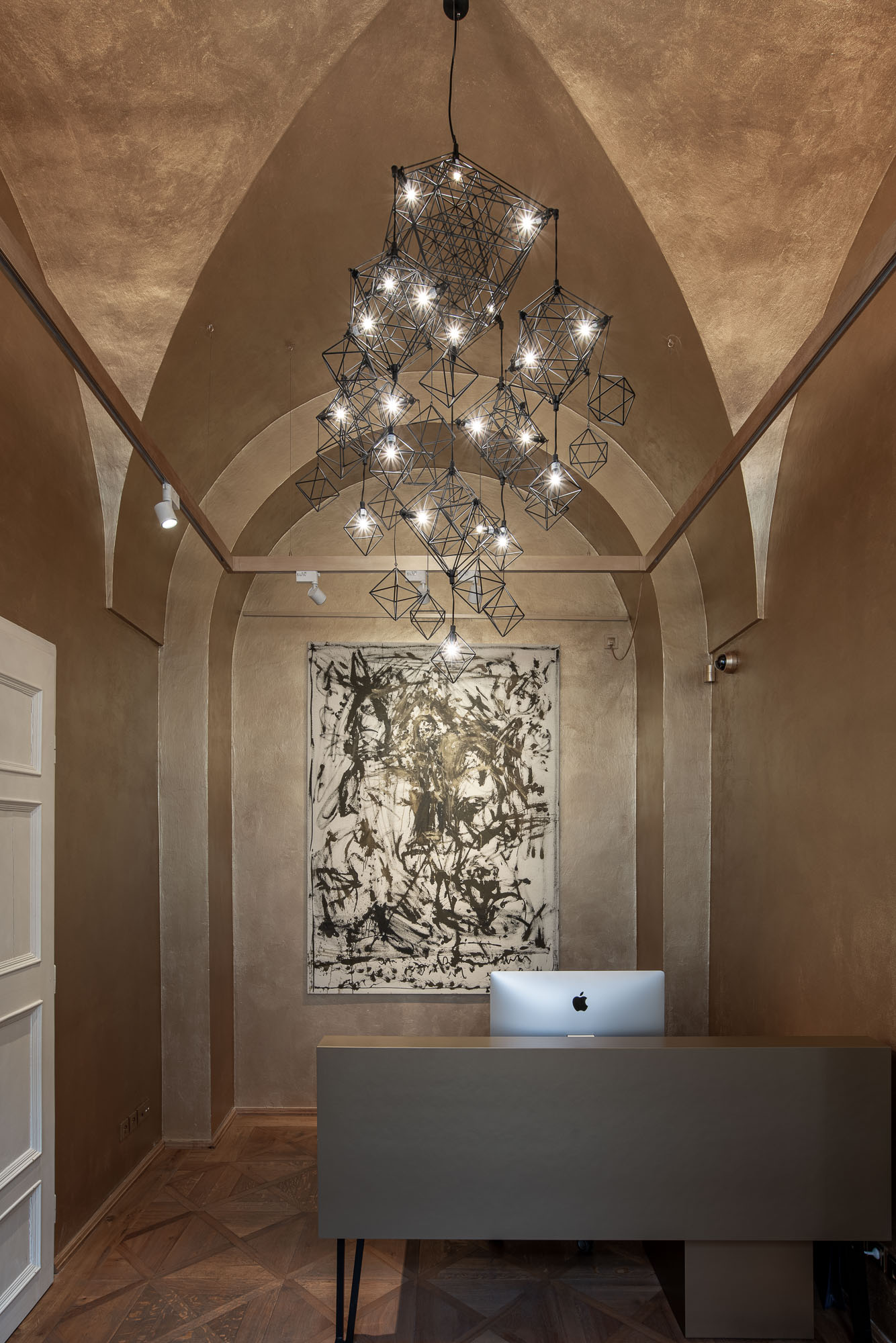 Lomený oblouk recepce galerie ARCIMBOLDO rámuje kaskádu drátěného svítidla nad recepcí s abstraktní malbou. Praha, Malá Strana.