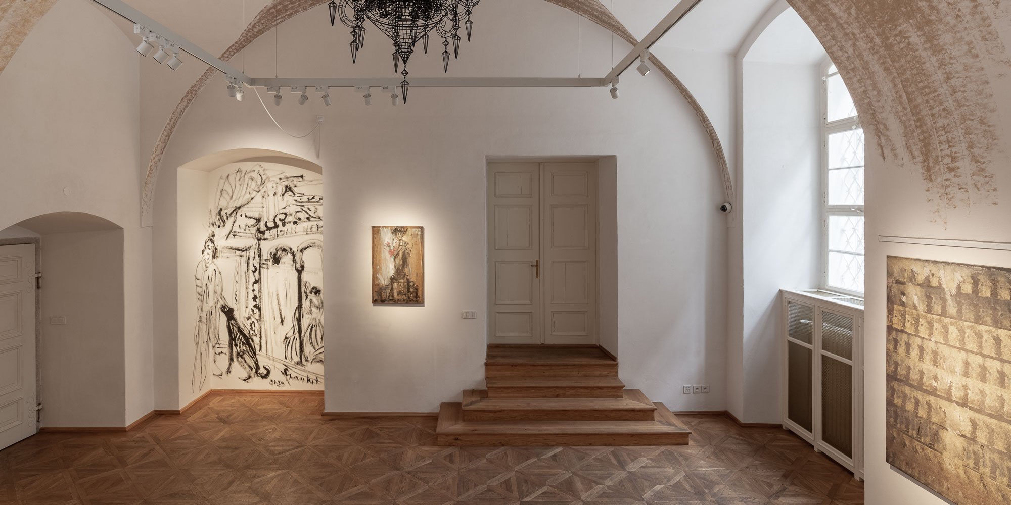 Průchozí prostor galerie ARCIMBOLDO s velkou figurální kresbou v klenuté nice, díly na stěnách a dřevěnými schody. Malá Strana, Praha.