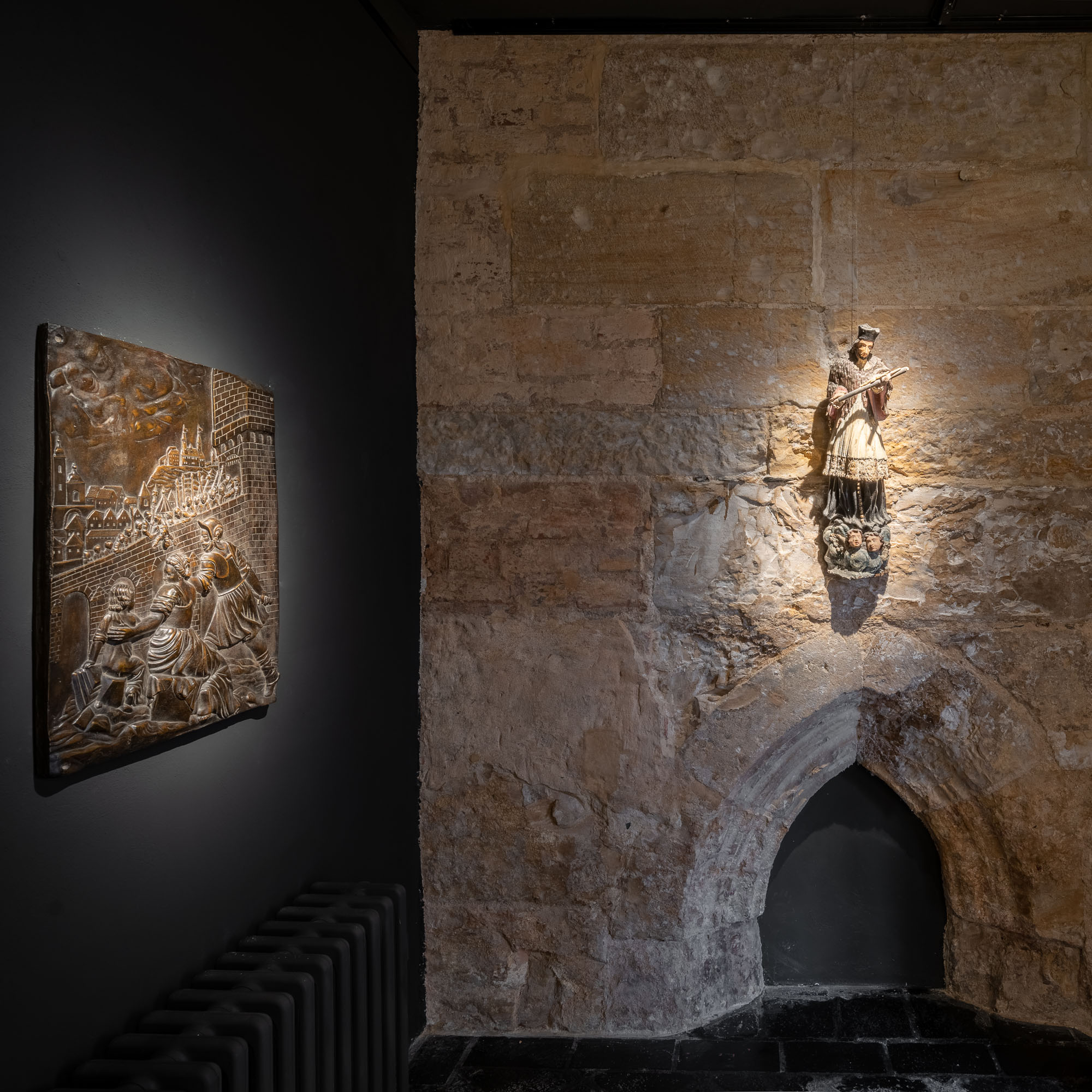 Temná výstavní místnost galerie ARCIMBOLDO s původní kamennou zdí, gotickou nikou, osvětlenou figurínou a bronzovým reliéfem. Praha.