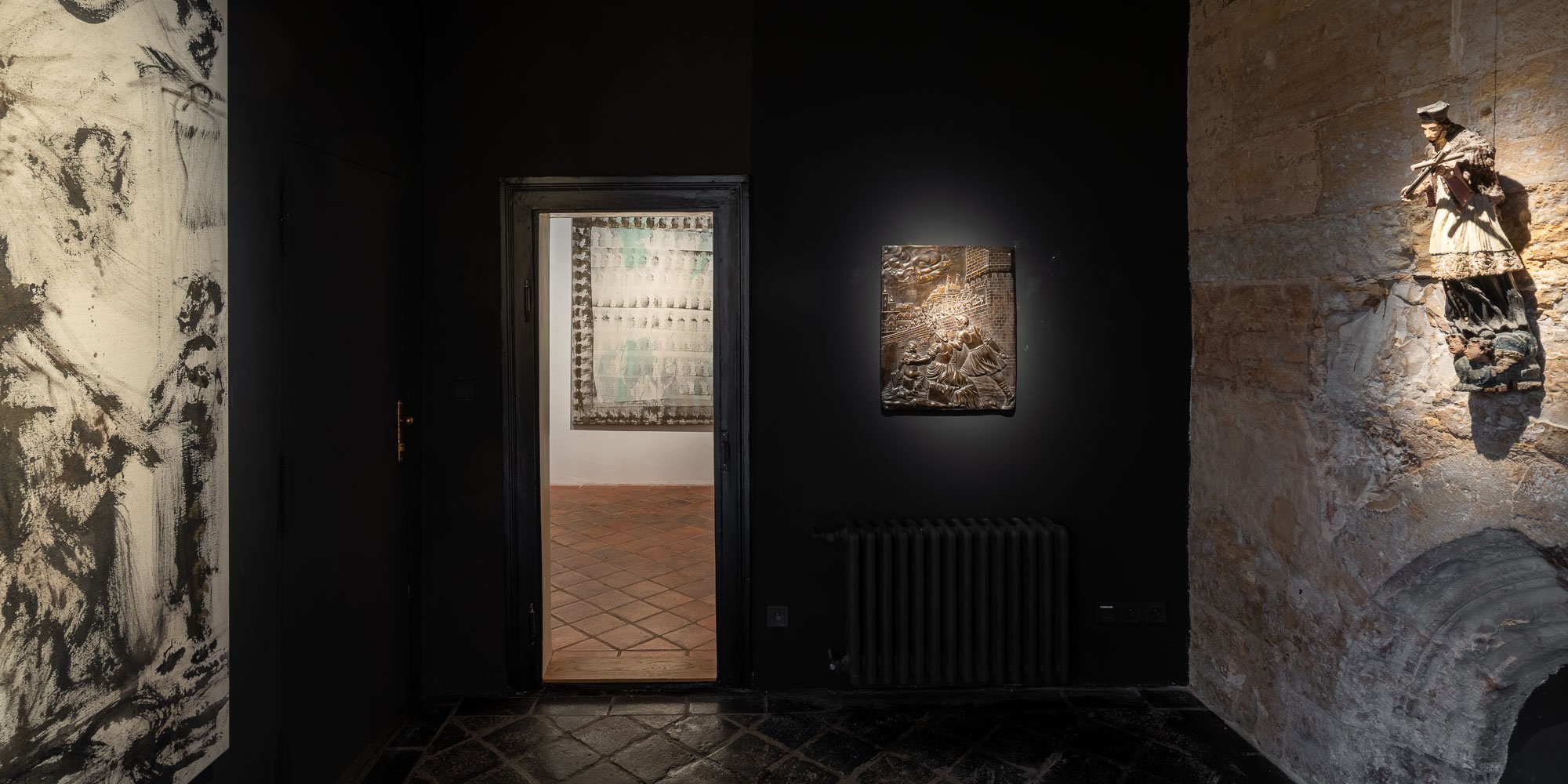 Panoramatický pohled na temnou místnost galerie ARCIMBOLDO s černými stěnami, gotickou nikou, figurínou, reliéfem a průchodem do galerie.