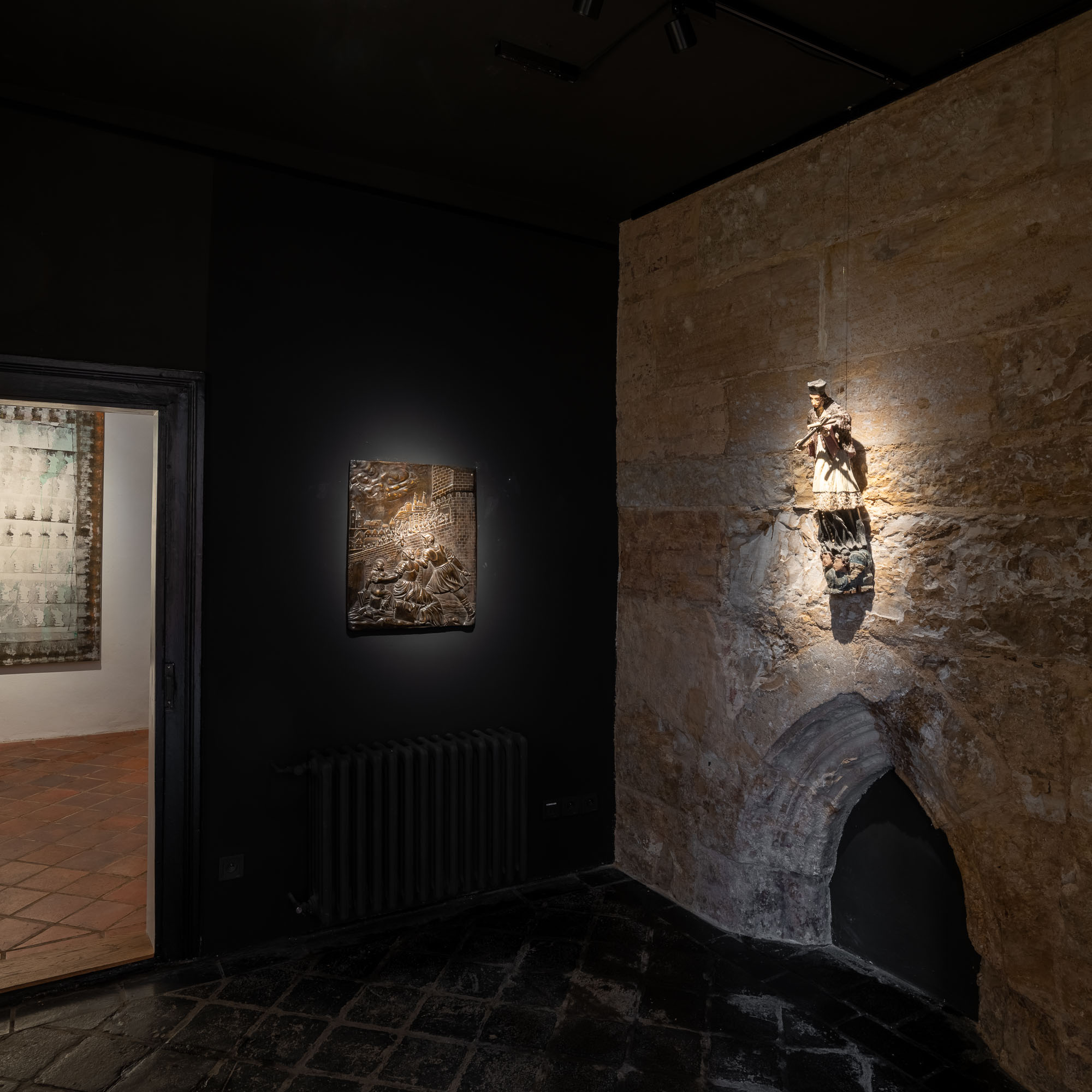 Panoramatický pohled na temnou místnost galerie ARCIMBOLDO s černými stěnami, gotickou nikou, figurínou, reliéfem a průchodem do galerie.