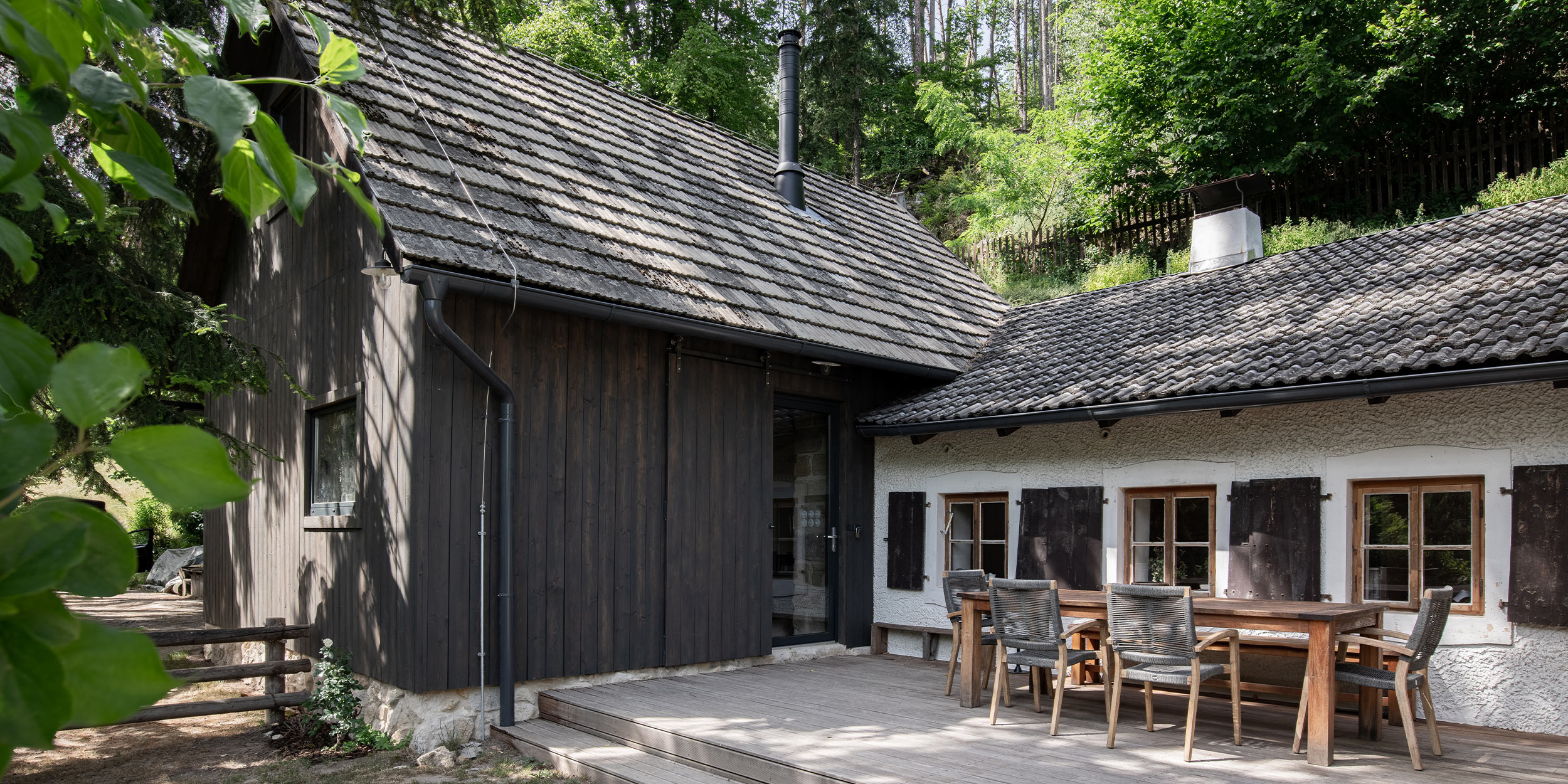Rekonstruovaná stodola Kokořín a přilehlá chalupa — sdílená dřevěná terasa s tíkovým zahradním stolem a židlemi mezi oběma objekty, černý kouřovod stodoly a stromy v pozadí