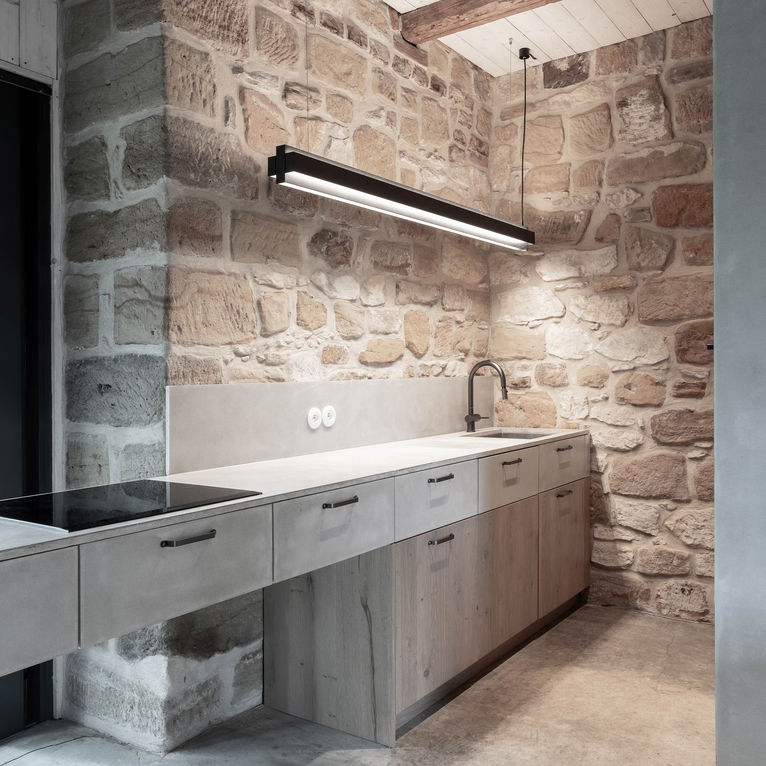 Side view of Kokořín barn kitchen counter — concrete worktop with black induction hob and concrete backsplash, linear LED pendant, oak kitchen cabinets in sandstone corner