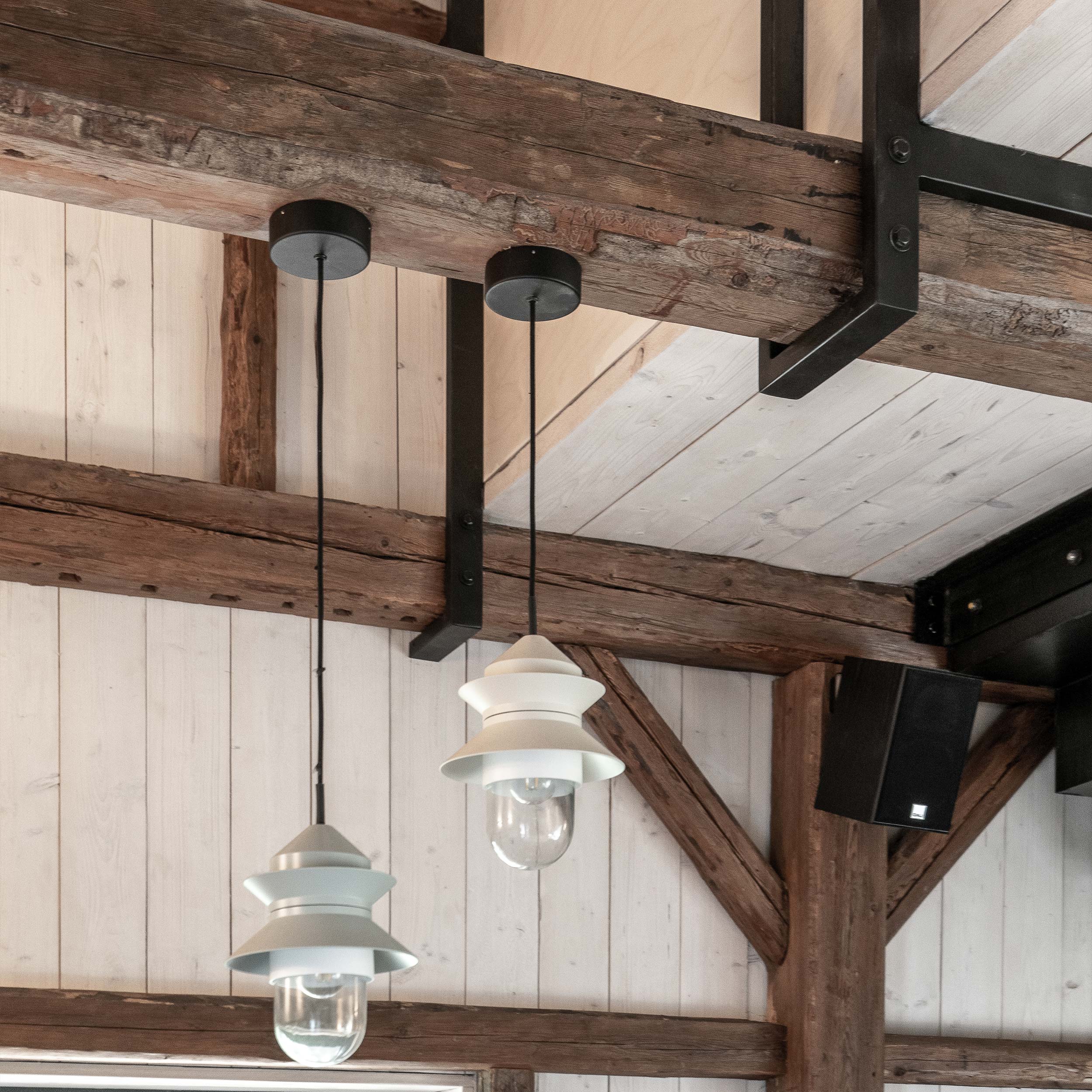 Dvě průmyslové závěsné lampy se světlým smaltovaným stínítkem a odhalenou žárovkou zavěšené z původní dřevěné trámové konstrukce — bílé palubkové stropy a černý ocelový spojovací prvek trám–ocel