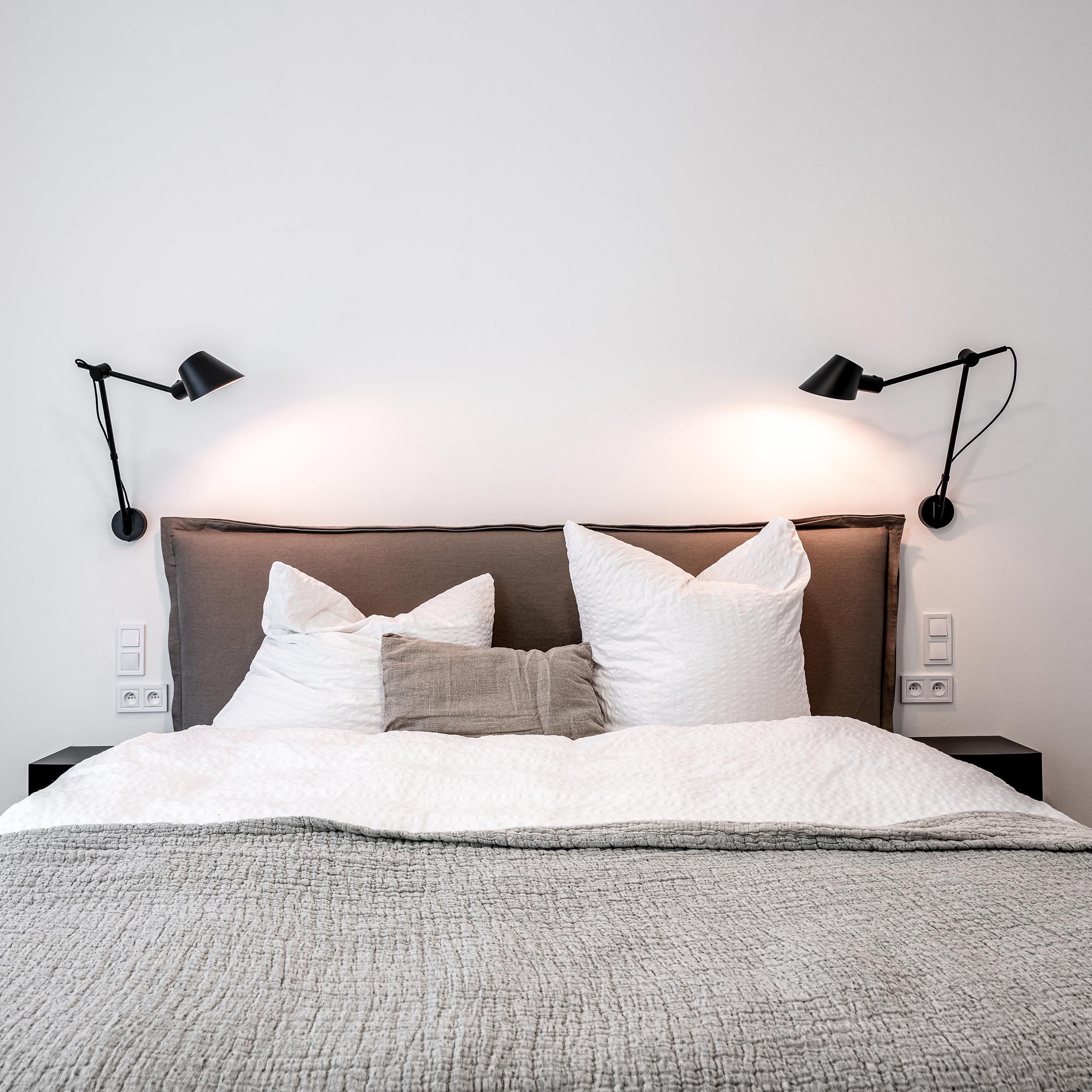 Čelní pohled na čalouněné čelo postele s bílým ložním prádlem, šedými polštáři a párem černých kloubových čtecích lamp – ložnice byt Jiřího Z Poděbrad OOOOX