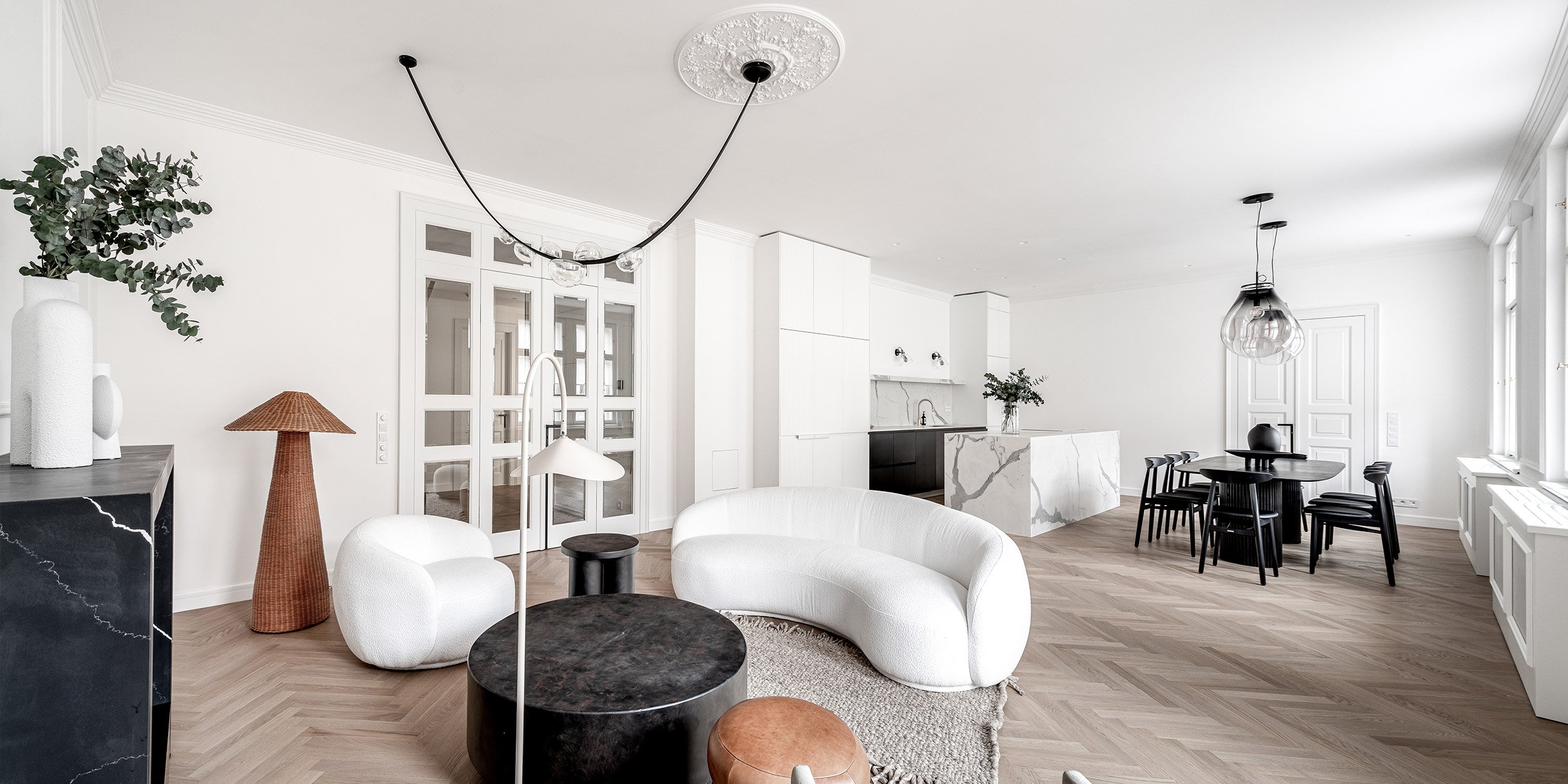 Panoramatický záběr celého apartmánu z krbu – krb, zaoblená bílá sedačka, kuchyňský ostrůvek, jídelní stůl, prosklené dveře a rybinová podlaha – vinohradský byt OOOOX