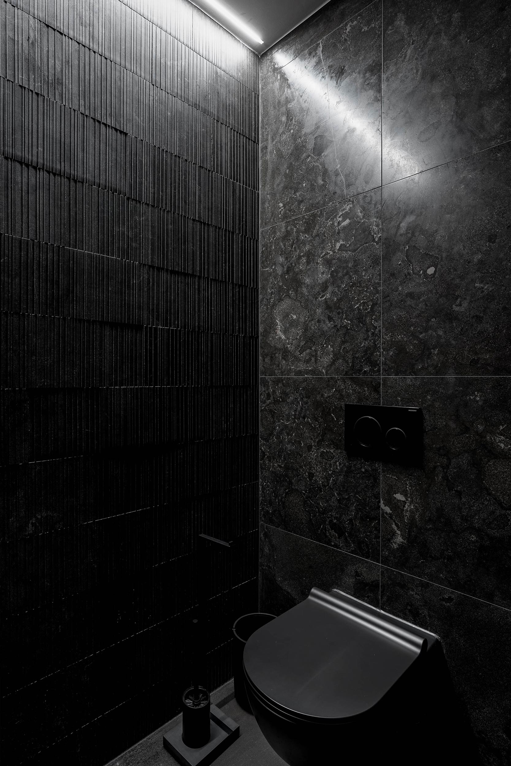 Tmavé WC butiku LA TABLE — černá závěsná mísa na pozadí tmavých strukturovaných obkladů a kamenné stěny, lineární světlo u stropu