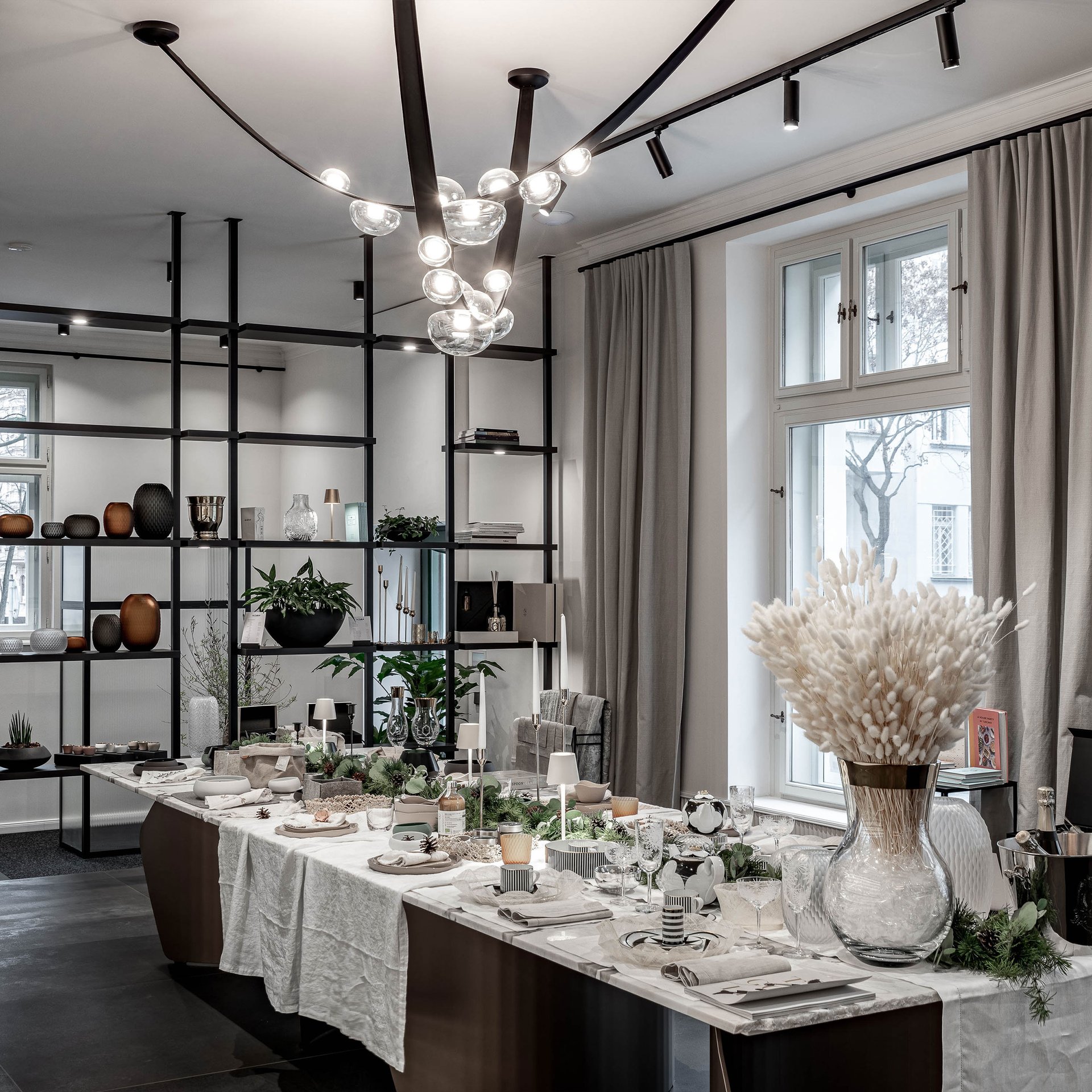 Showroom LA TABLE s vánočním stylingem — kvartzitový stůl s dekorativními suchými květinami, chvojím a svíčkami; ocelový regálový systém vlevo; velká okna; organické stropní světlo