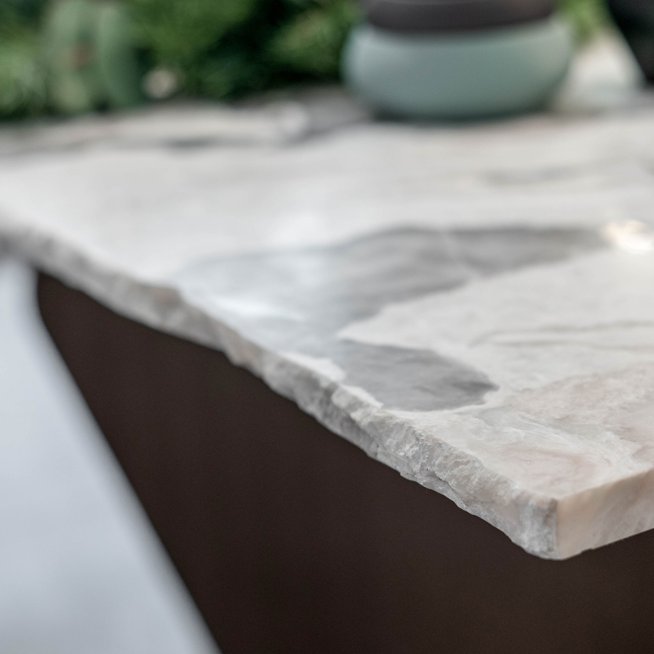 Makro detail hrany kvartzitového stolu LA TABLE — surová štípaná lomená hrana odhalující přirozenou strukturu kamene, tmavý ocelový podstavec stolu
