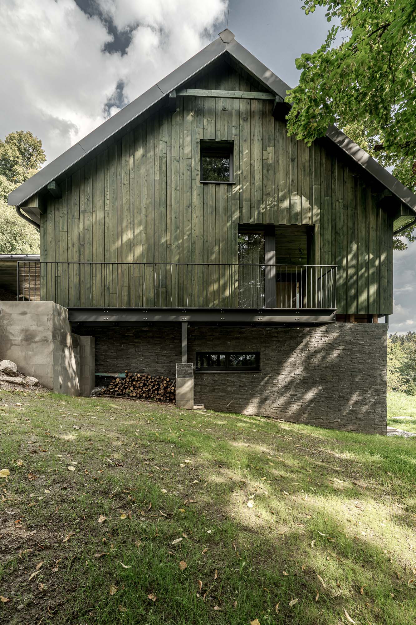 Zadní exteriér horské chalupy MARIÁNSKÁ – zelená svislá dřevěná fasáda, tmavá sedlová střecha, kamenný a cihlový sokl se zásobou dřeva a balkon s kovovým zábradlím