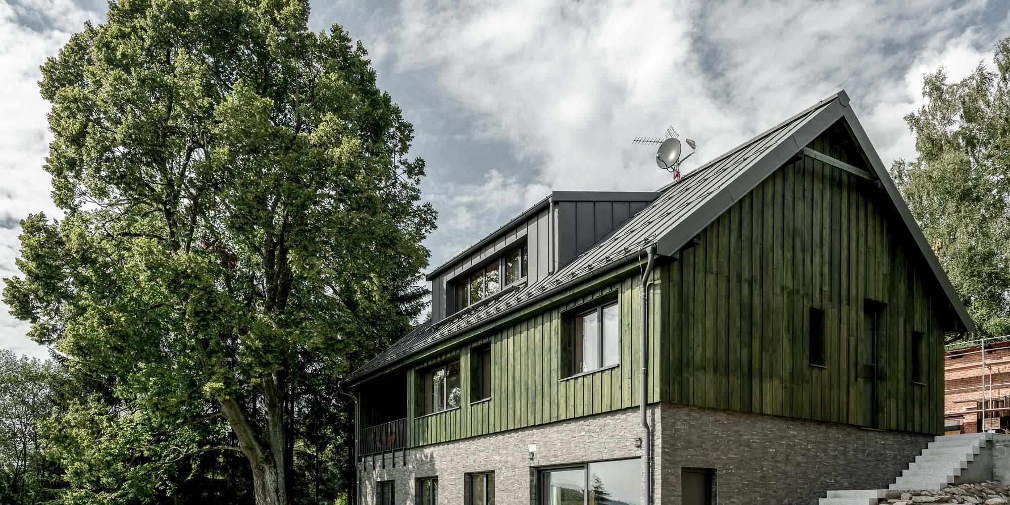 Boční pohled na exteriér chalupy MARIÁNSKÁ – zelená dřevěná fasáda, tmavá střecha, kamenný a cihlový sokl s balkonem a velký strom v popředí