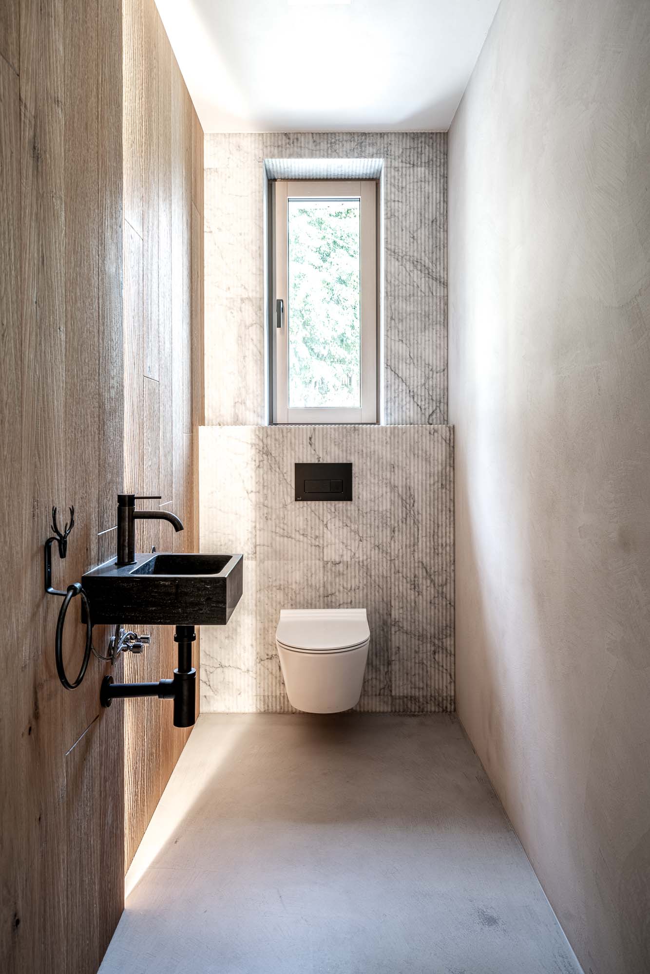 Designová toaleta horské chalupy MARIÁNSKÁ – úzká místnost se žlábkovaným mramorovým obkladem, závěsnou toaletou, malým černým kamenným umyvadlem a dřevěnou stěnou