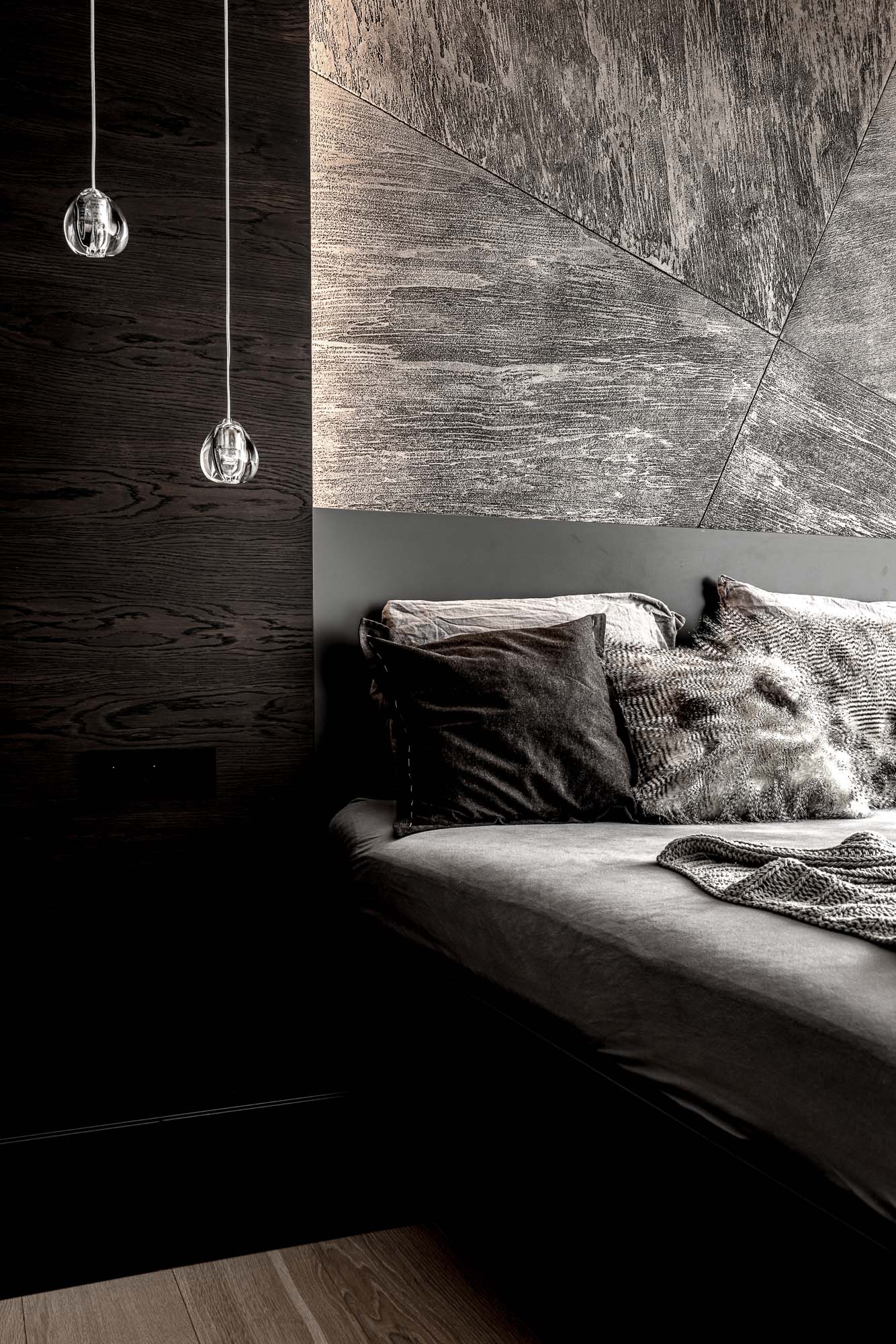 Bedroom side detail in the Černošice house – dark plaster headwall and dark wood vertical panel with crystal pendant lights, grey bed with dark and fur cushions