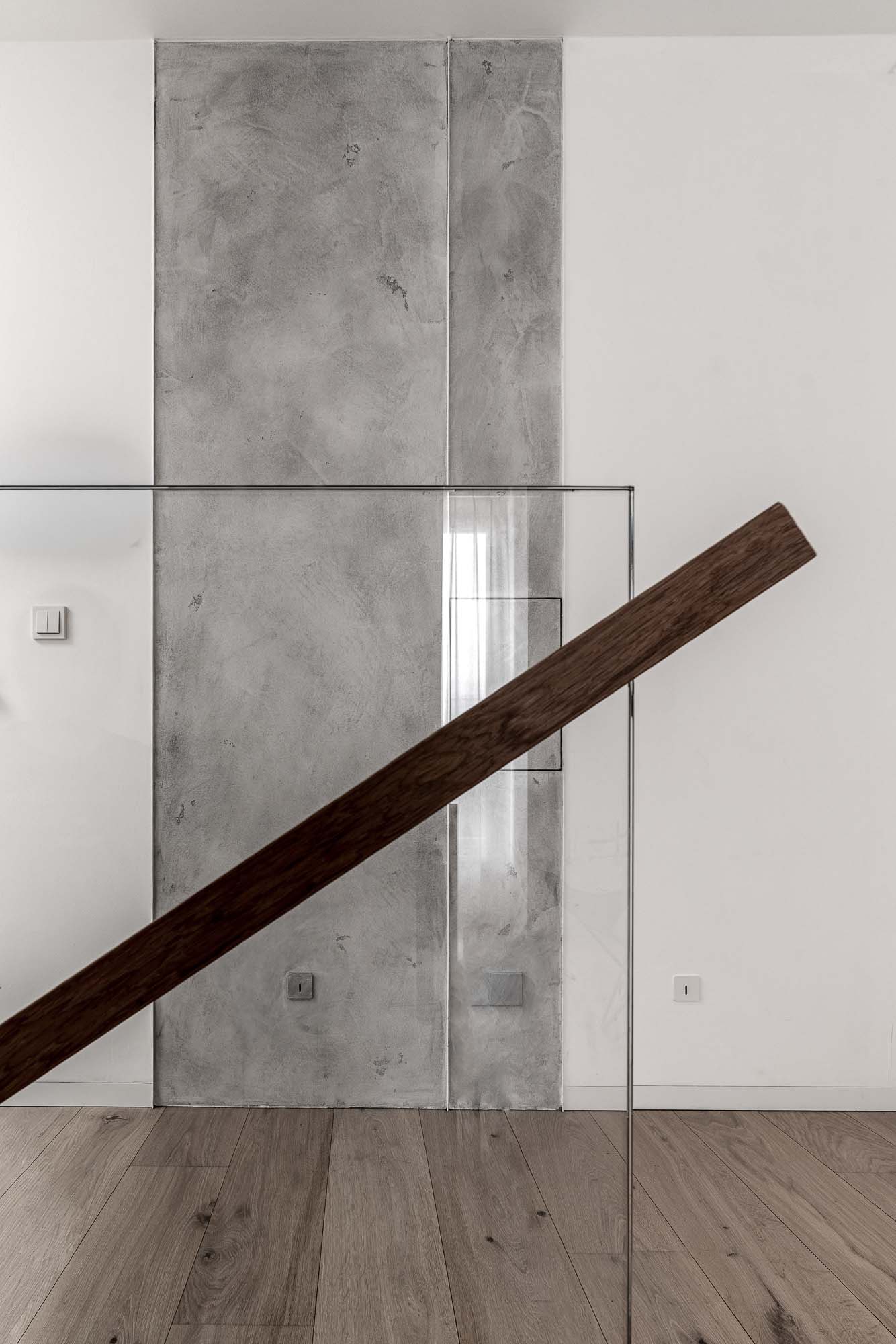 Detail schodiště domu Černošice – skleněné zábradlí s tmavým dřevěným madlem, stěrkové panely v pozadí, světlá dubová podlaha