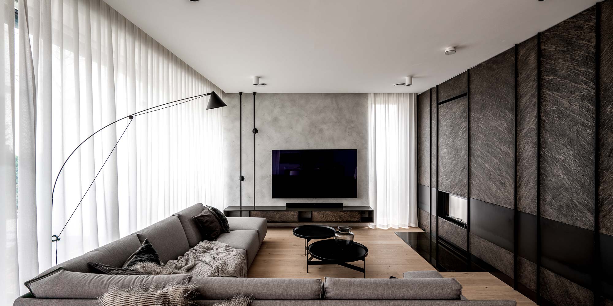 Panoramatický pohled na obývací pokoj domu Černošice – šedá modulová pohovka, černá oblouková lampa, TV na šedé stěně, tmavá krbová stěna vpravo, bílé závěsy, přirozené světlo