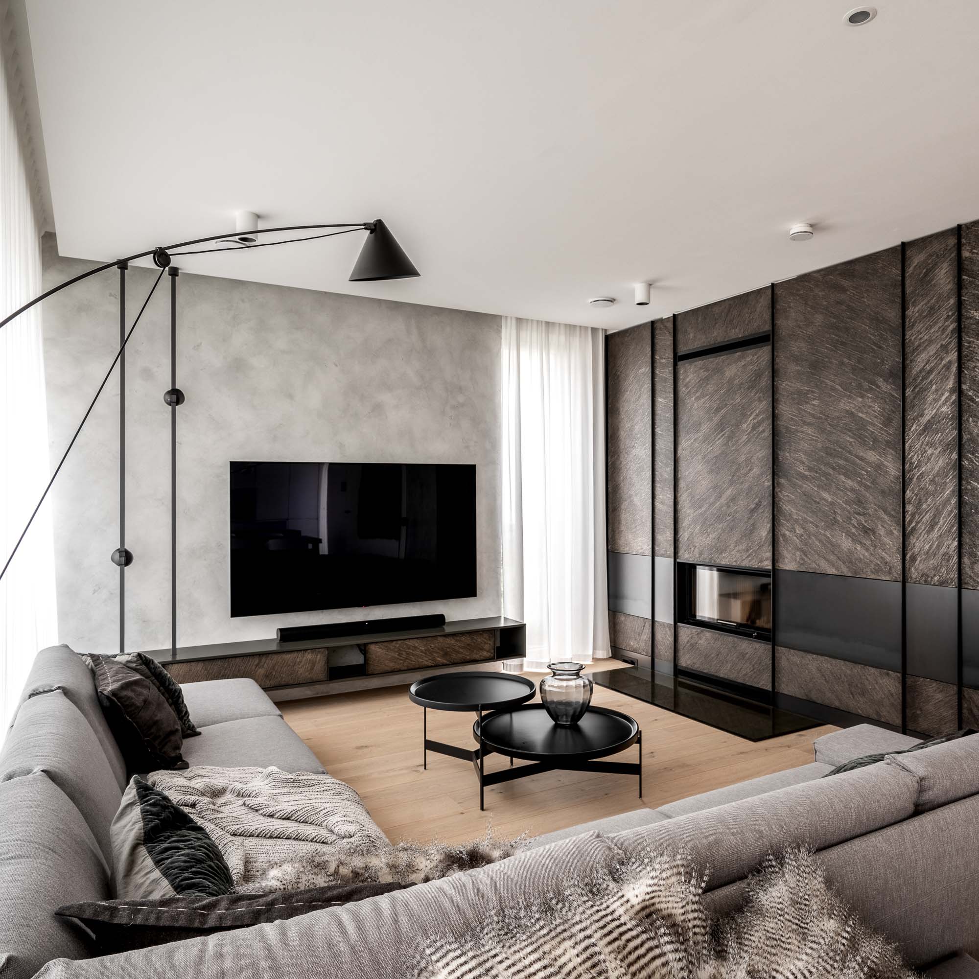 Obývací pokoj domu Černošice – šedá rohová pohovka, dva kulaté černé stolky, černá oblouková lampa, TV na šedé stěrkové stěně, krbová stěna s tmavou stěrkou a krb