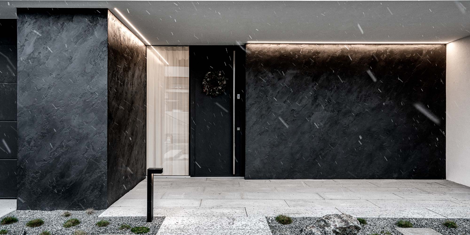 Entrance facade of the Černošice house in snowfall – dark textured plaster on the facade, black entrance door with decorative wreath and LED strip light, concrete path with gravel and rocks