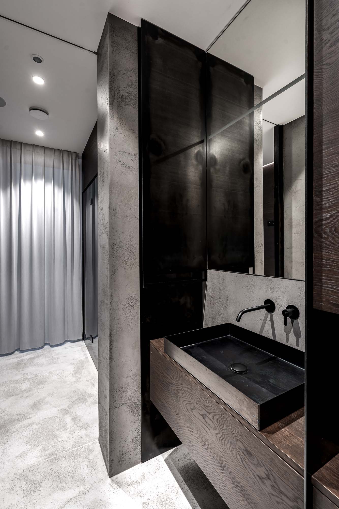Wellness koupelna domu Černošice – tmavé stěrkové stěny, tmavý dřevěný vanity s černým kamenným umyvadlem, nástěnné černé armatury, velké zrcadlo, tmavě lakovaná skříň