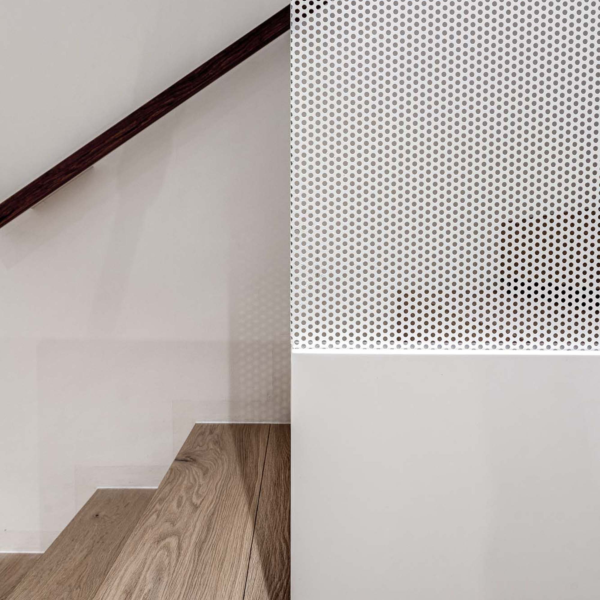 Detail schodiště domu Černošice – tmavé dřevěné madlo a stupeň schodiště, bílá stěna, perforovaný bílý kovový panel v detailním záběru