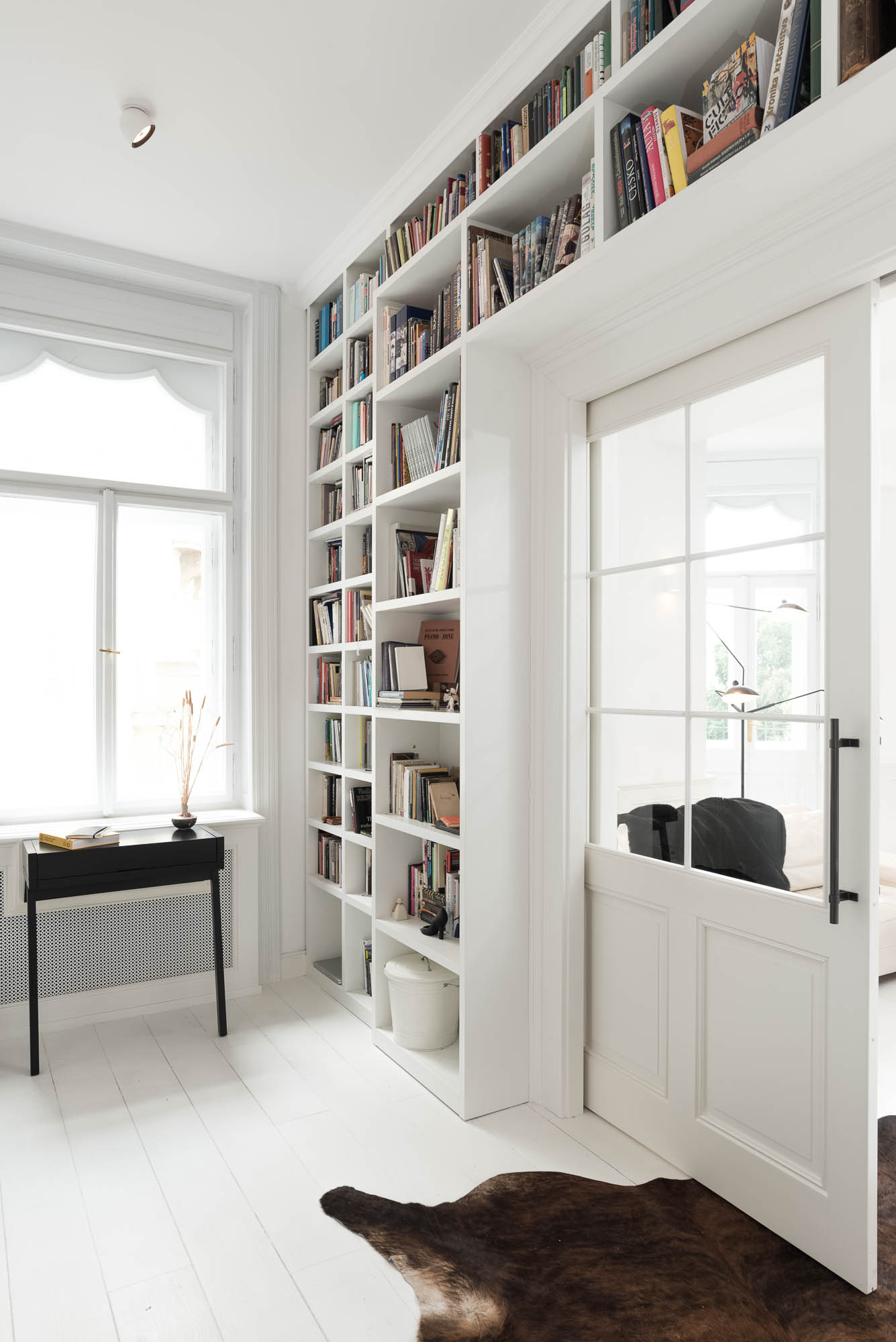 Study with a full-height bookshelf connected to the living area through pocket sliding doors, white wooden flooring | design by OOOOX