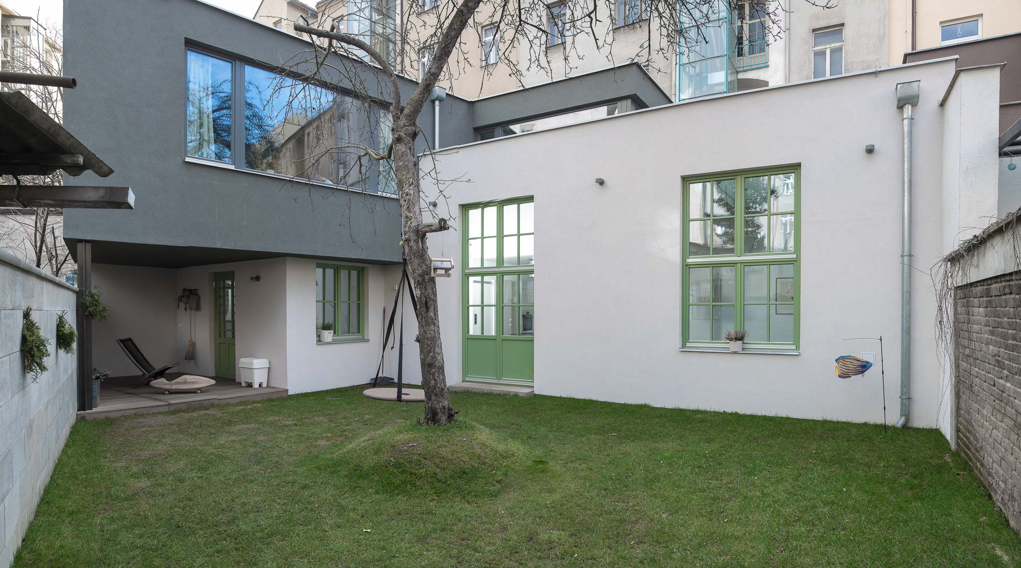 The courtyard garden with a lawn, tree with a swing, yellow stools, and the white rendered facade of the original studio with its green-framed windows; the grey new addition rises behind.