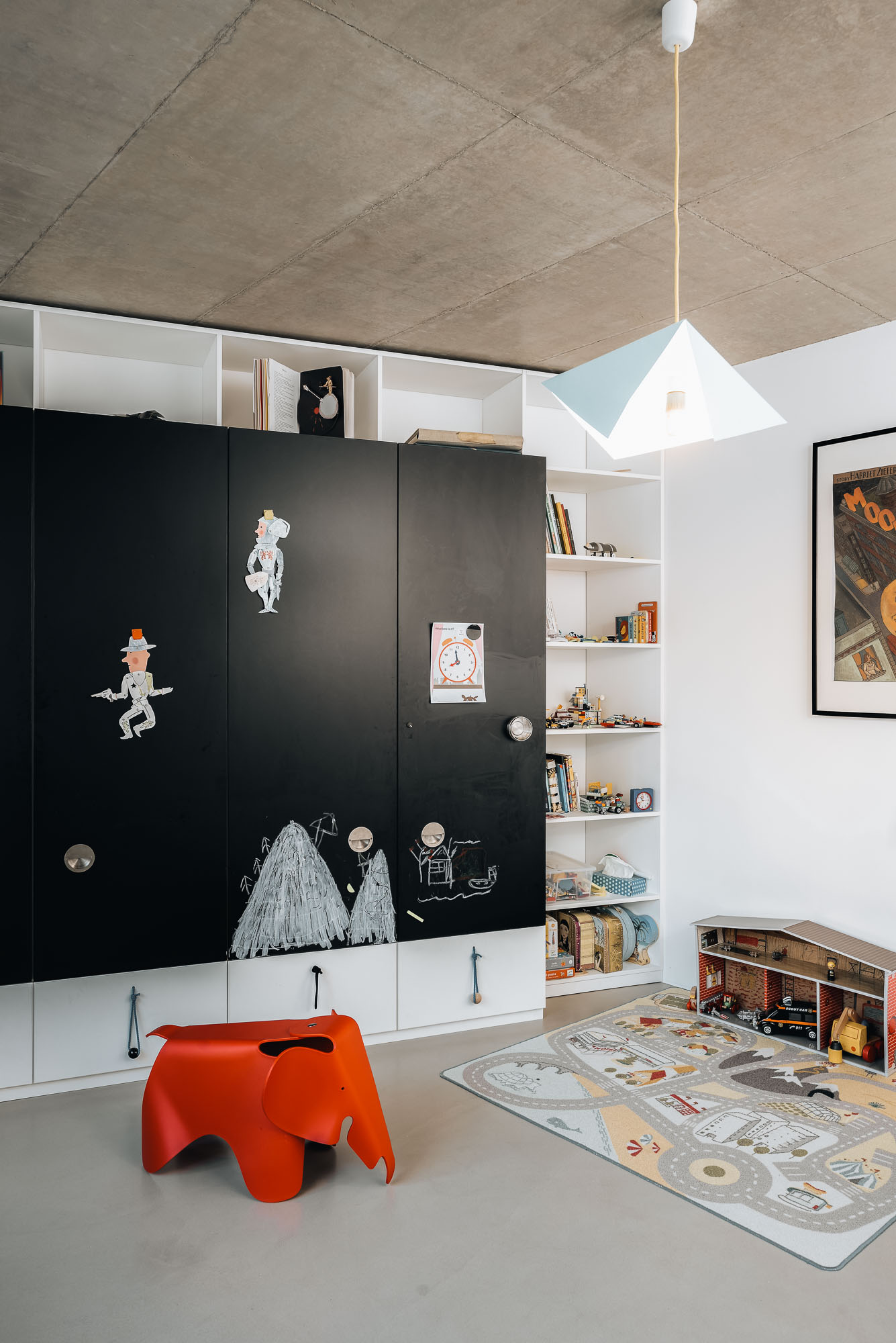 Dětský pokoj v novém nadzemním objemu s černou skříní s tabulovou plochou posetou křídovými kresbami, oranžovým slonem Eames, hracím kobercem, bílými policemi a pohledovým betonovým stropem.