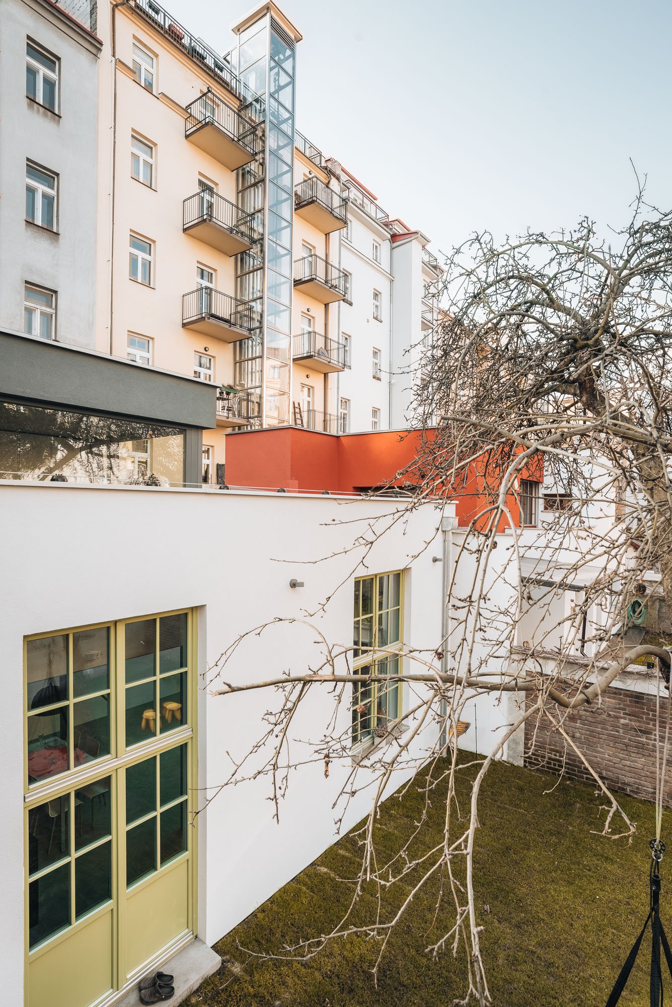 Exteriérový pohled z dvora na bílou omítnutou fasádu původního sochařského ateliéru se zelenými okny a šedým novým objemem viditelným nad střechou.