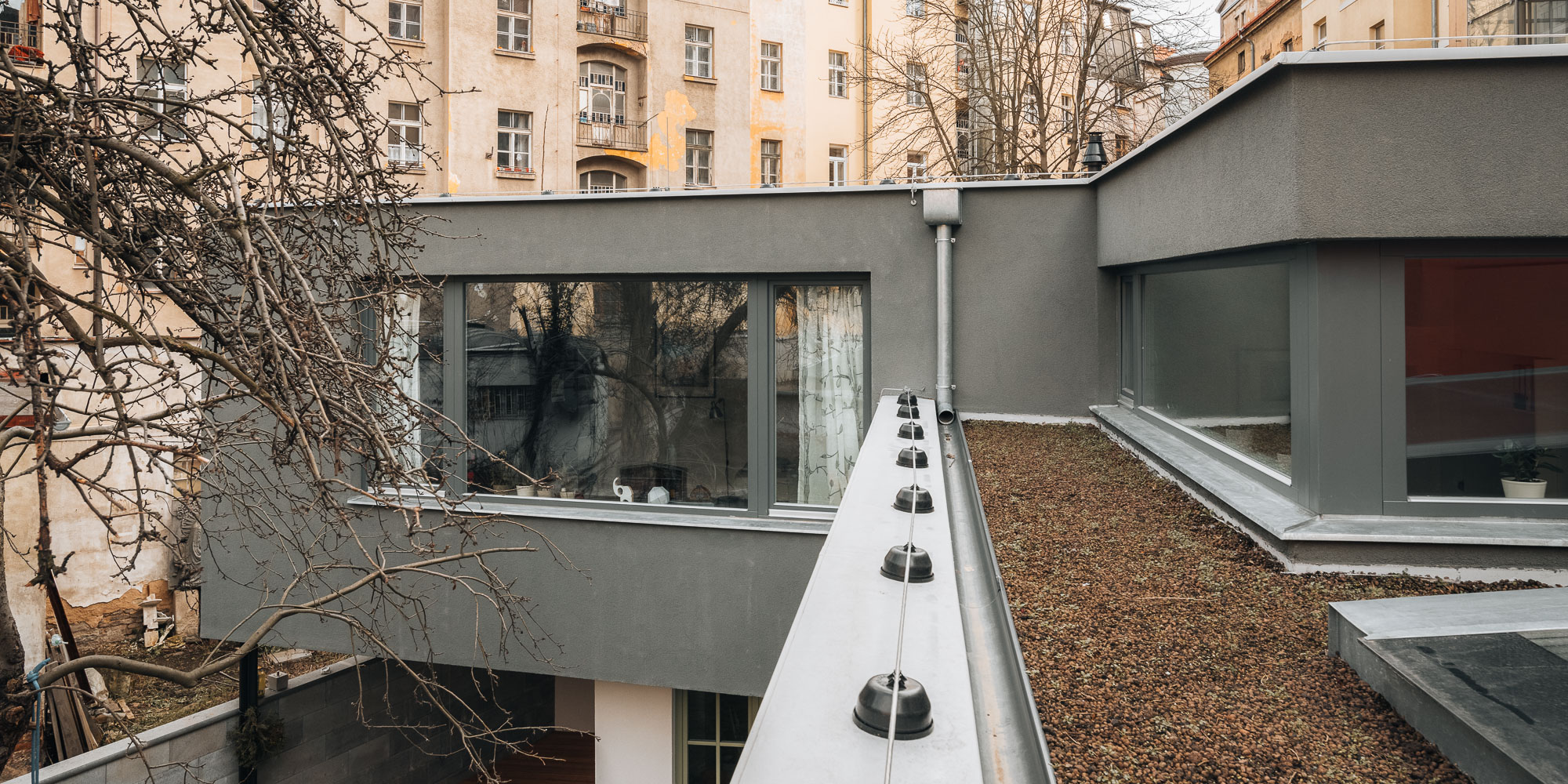View at roof level showing the gravel-covered roof garden between the new upper volume and the original studio roofline, with strip windows and the surrounding courtyard buildings.
