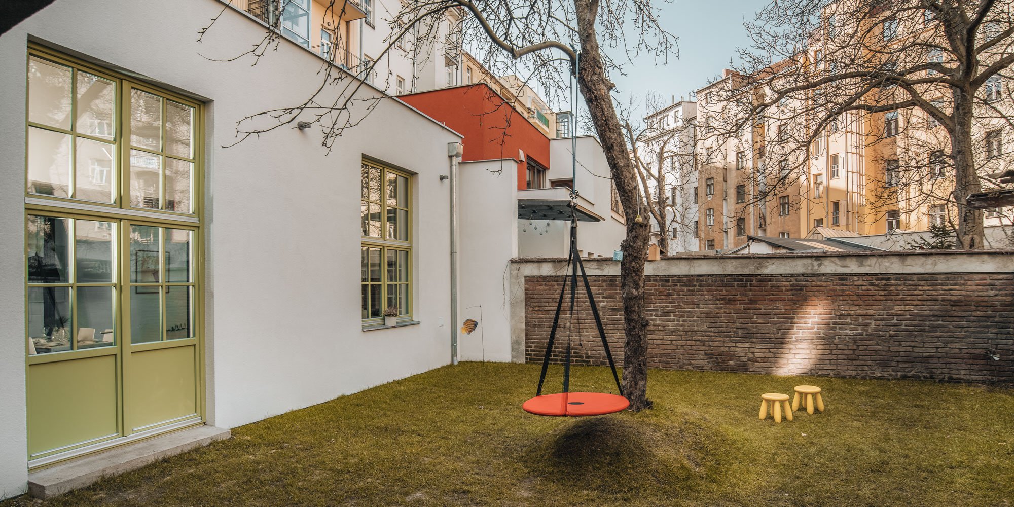 Celkový exteriérový pohled na zahradu ve dvoře a bílou fasádu původního ateliéru se zelenými okny, stromem s oranžovou kulatou houpačkou, žlutými stoličkami na trávě, cihlovou zahradní zdí