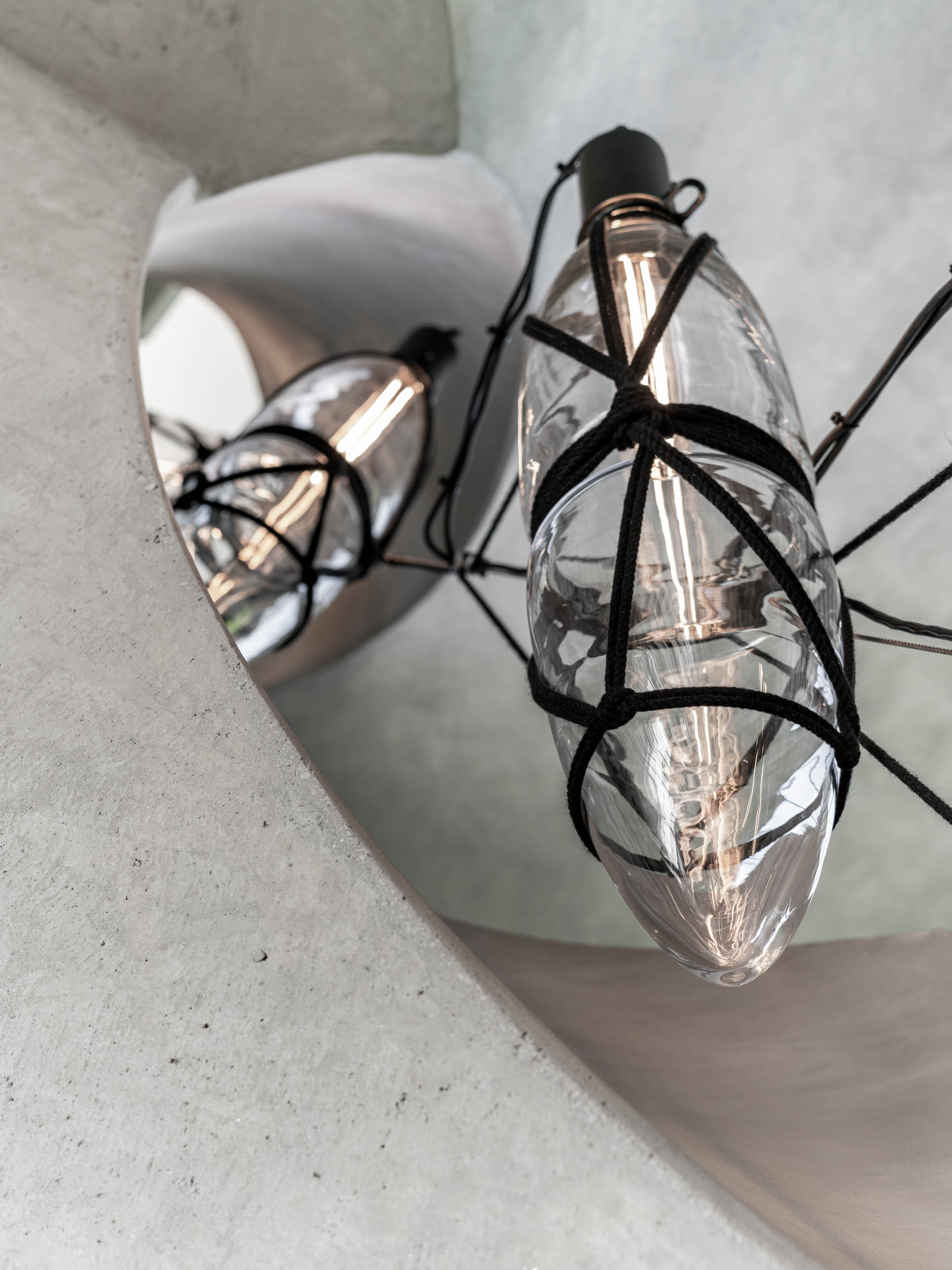 Makro detail závěsné lampy uvnitř schodiště vily Ohnivcova — skleněná kapková lampa svázaná černými šňůrami, odrazové světlo na pohledovém betonu středové šachty