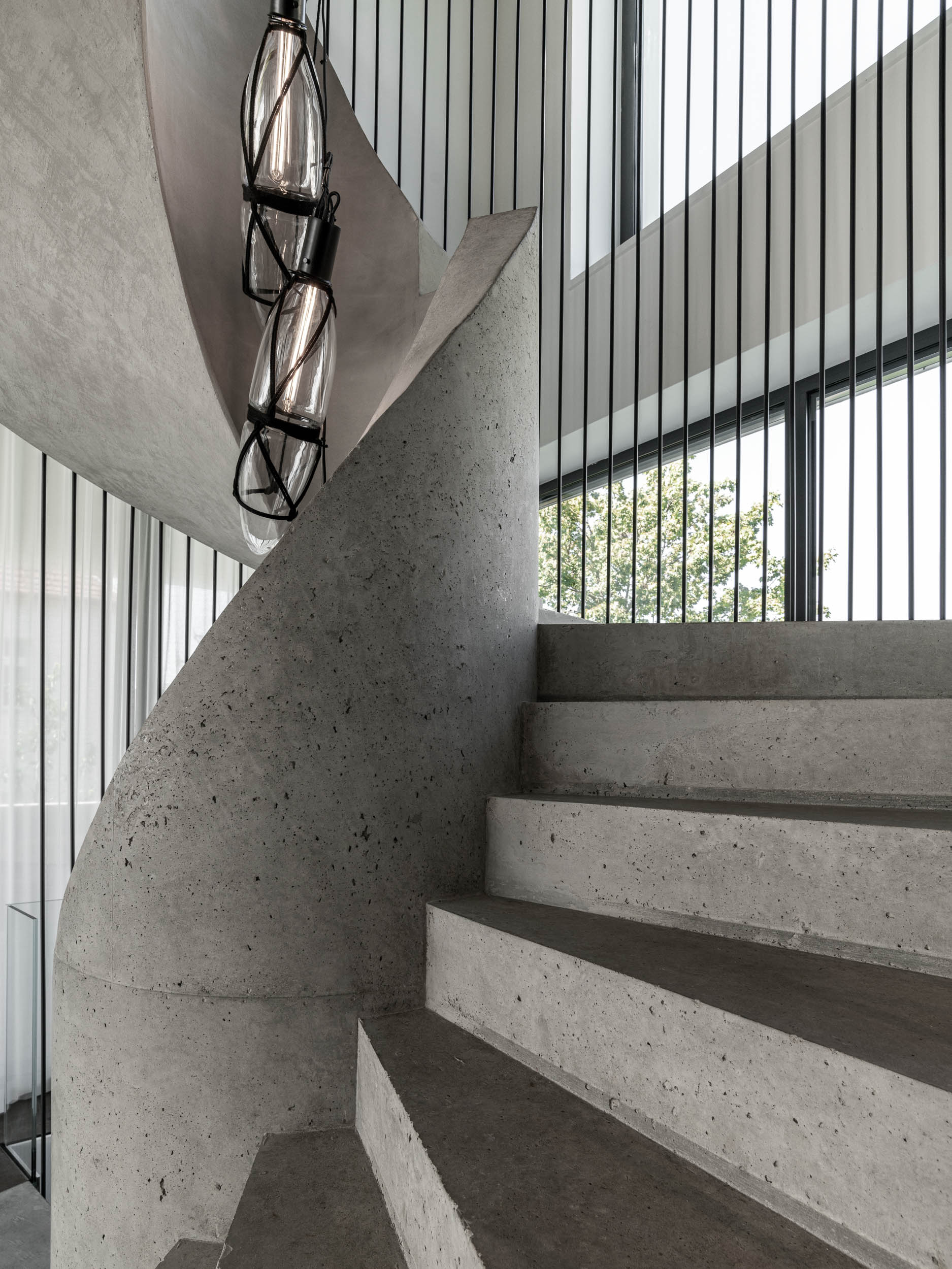 Side view of the concrete spiral staircase of Villa Ohnivcova — curved central core, exposed concrete steps, steel rod enclosure, teardrop pendants inside, view through the window to the garden