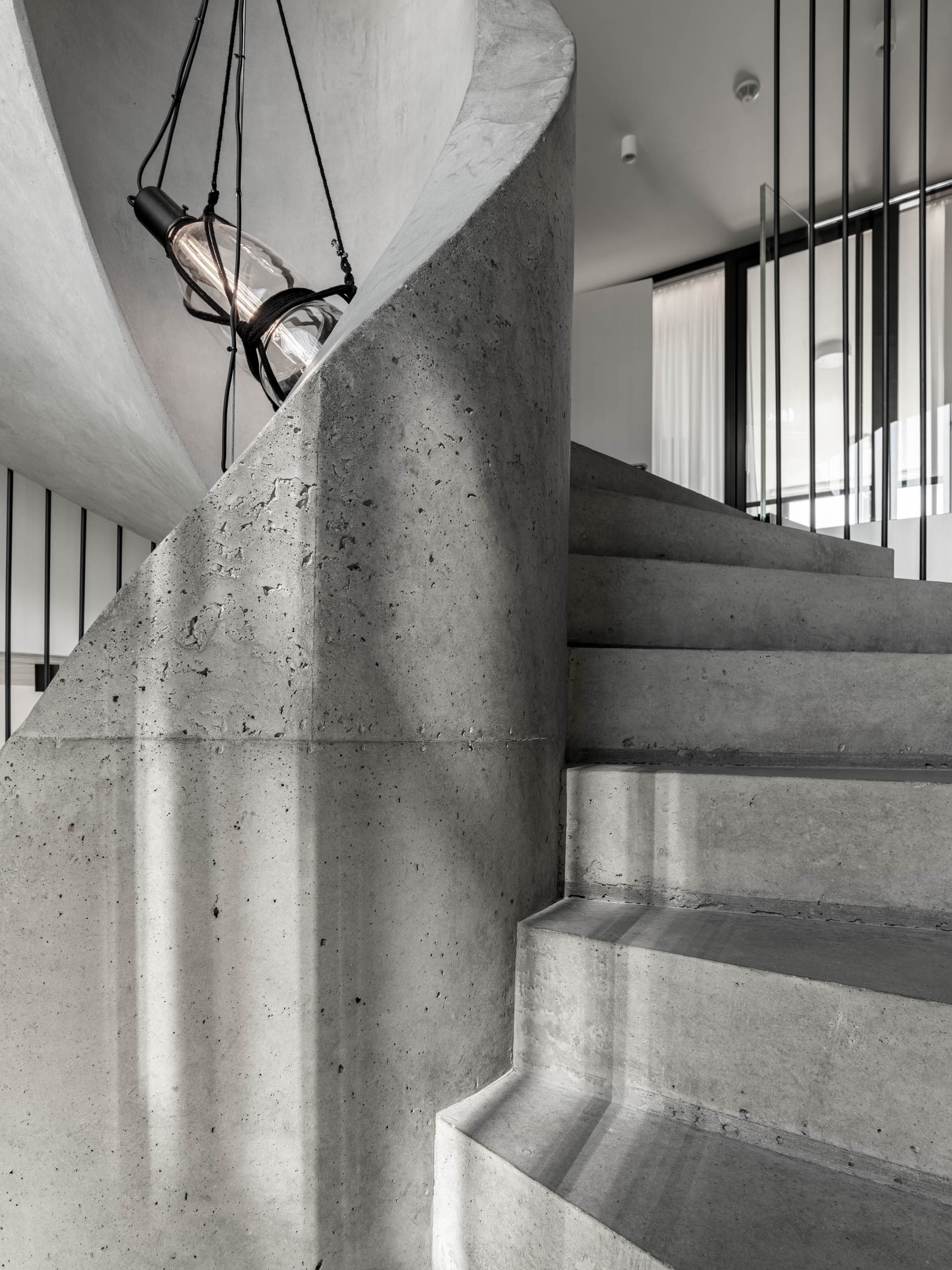 Detail točitého betonového schodiště vily Ohnivcova — oblý tvar středové šachty pohledového betonu, betonové stupně, kapkové svítidlo, ocelová tyč tubusového zábradlí v pozadí