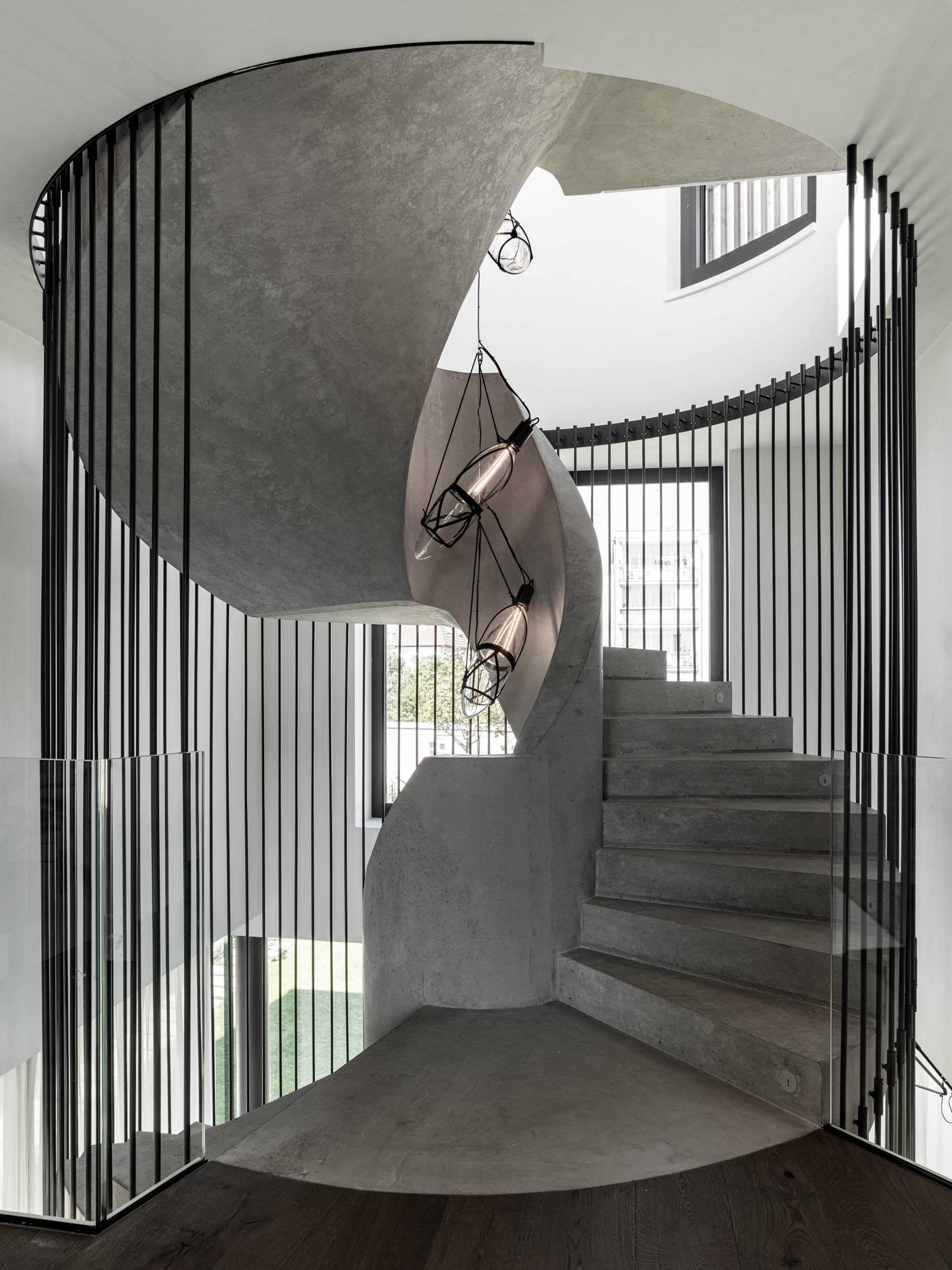 Full view of the concrete spiral staircase of Villa Ohnivcova from the ground floor — three-storey concrete spiral enclosed by a steel rod structure, glass railing at the base, suspended pendants, lit step edges
