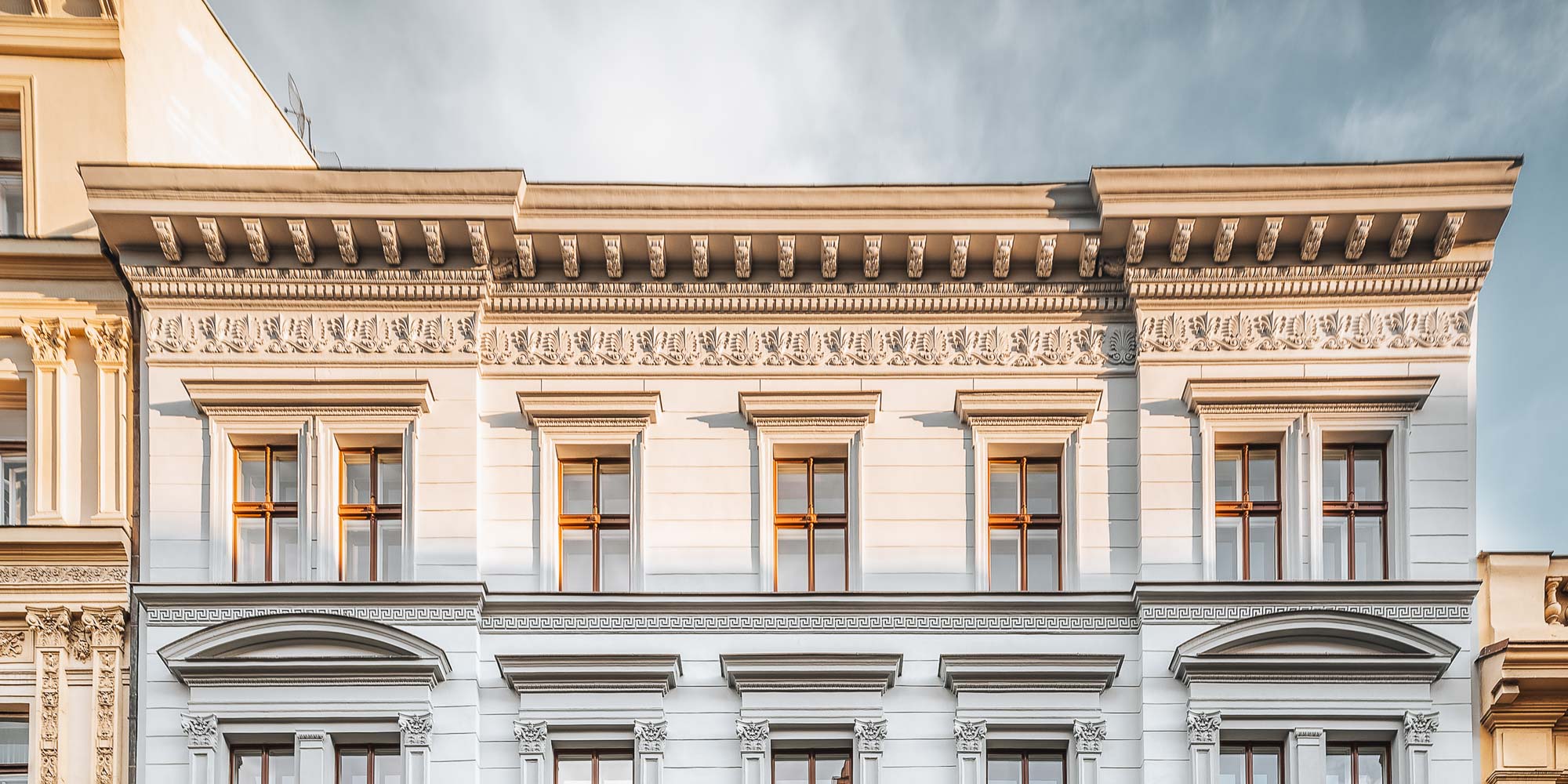 Detail horní části uliční fasády bytového domu Opletalova se štukovými římsami a akantovými pásy osvícenými odpoledním sluncem