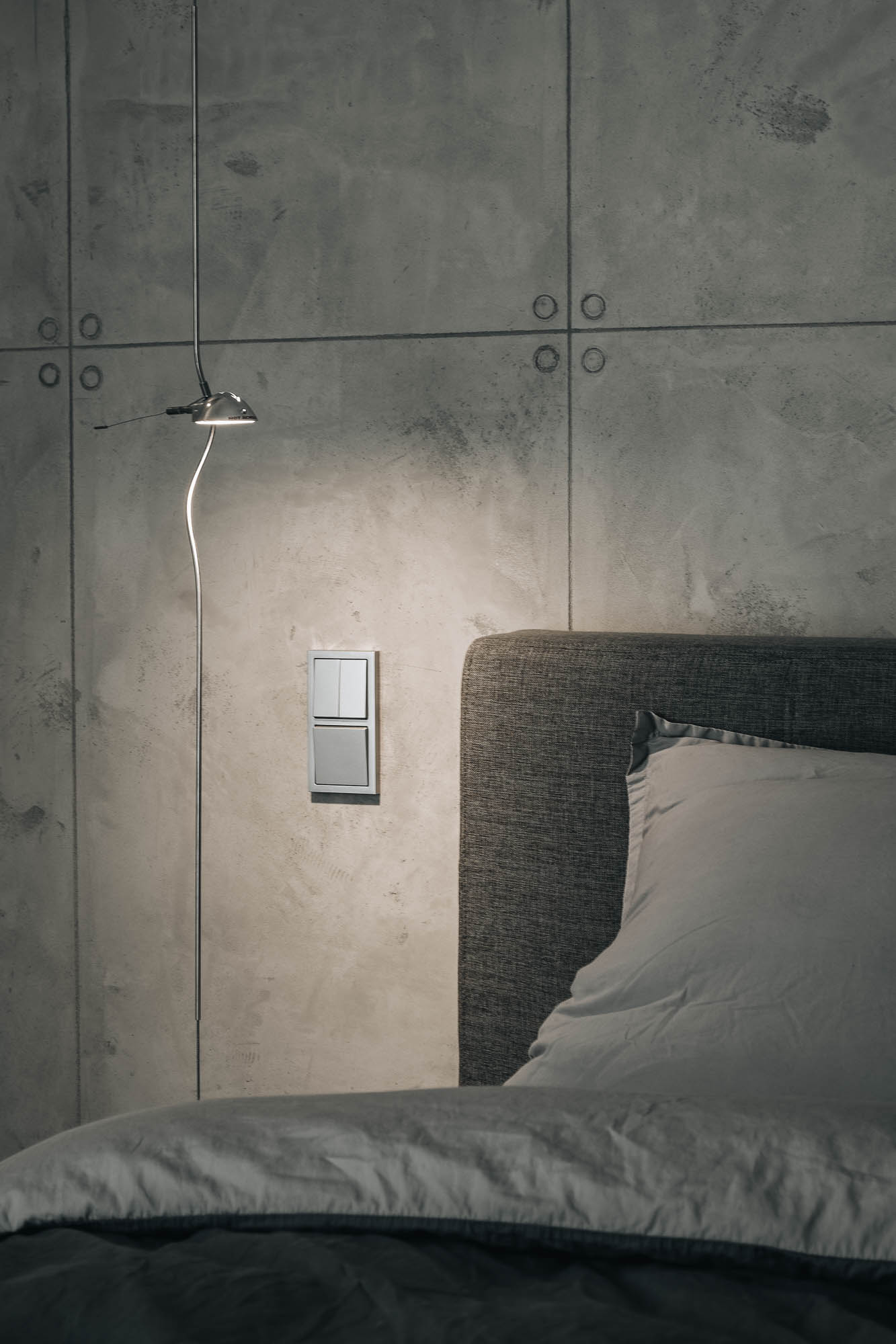 Detail nástěnného svítidla vedle čalouněné postele na pozadí velkoformátového pohledového betonu ve vile