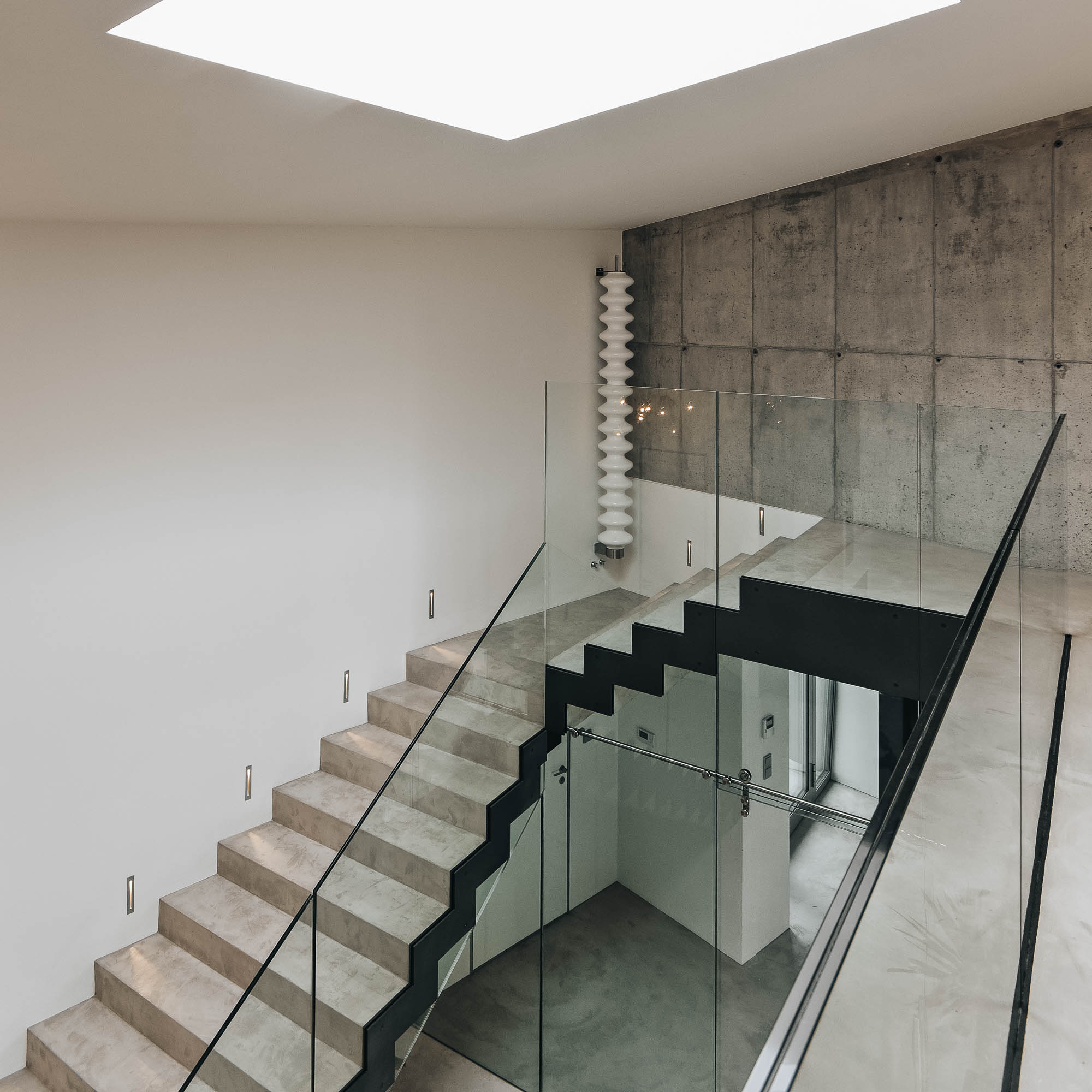 Schodiště vily s pohledovým betonem, skleněným zábradlím, designovým radiátorem a střešním světlíkem