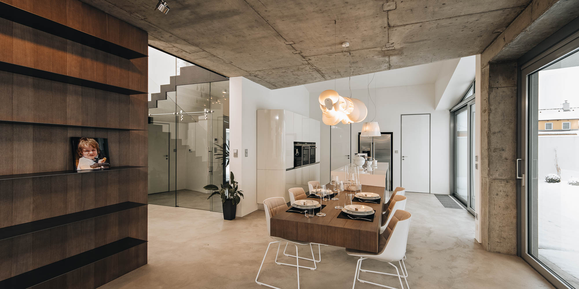 Jídelna vily s pohledovým betonovým stropem, vykonzolovaným stolem a dřevěnými policemi, schodiště v pozadí