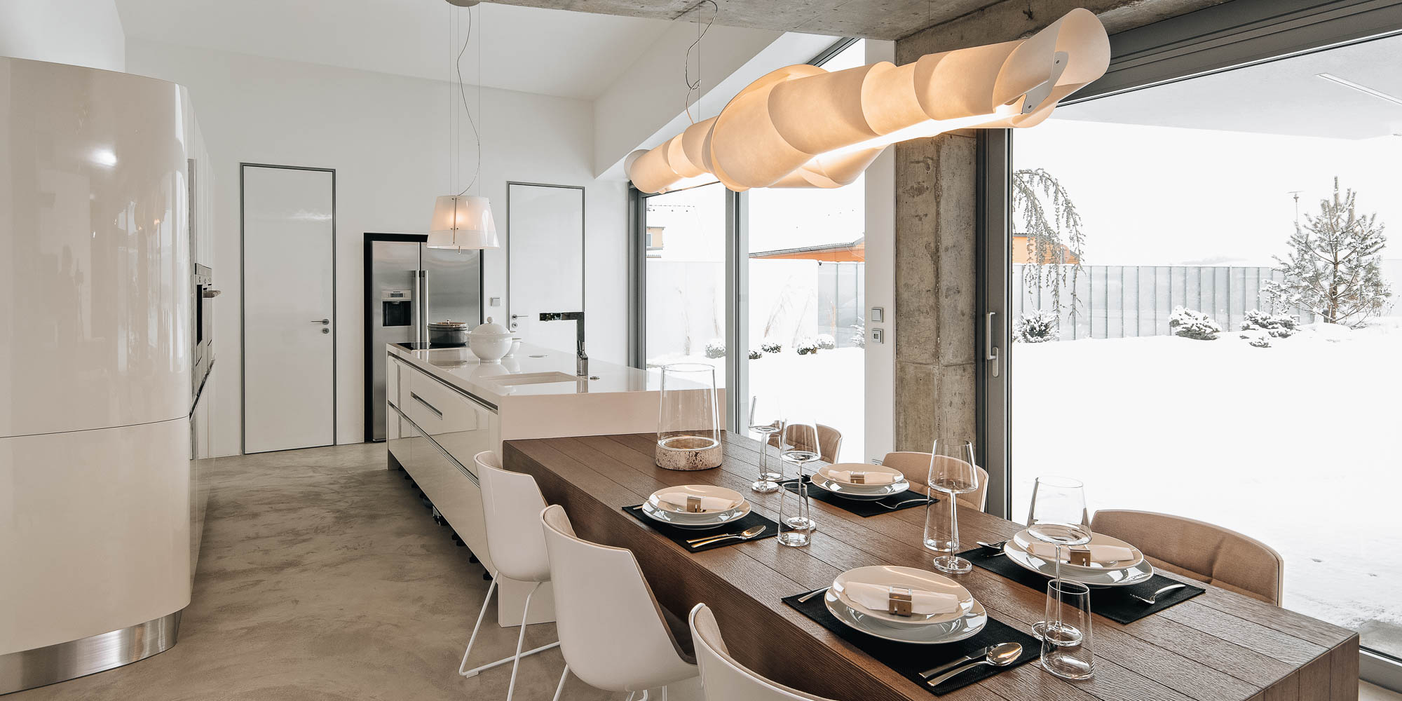 Kuchyně s bílými lesklými frontami a jídelna s designovým svítidlem a vykonzolovaným jídelním stolem v interiéru vily, zimní výhled