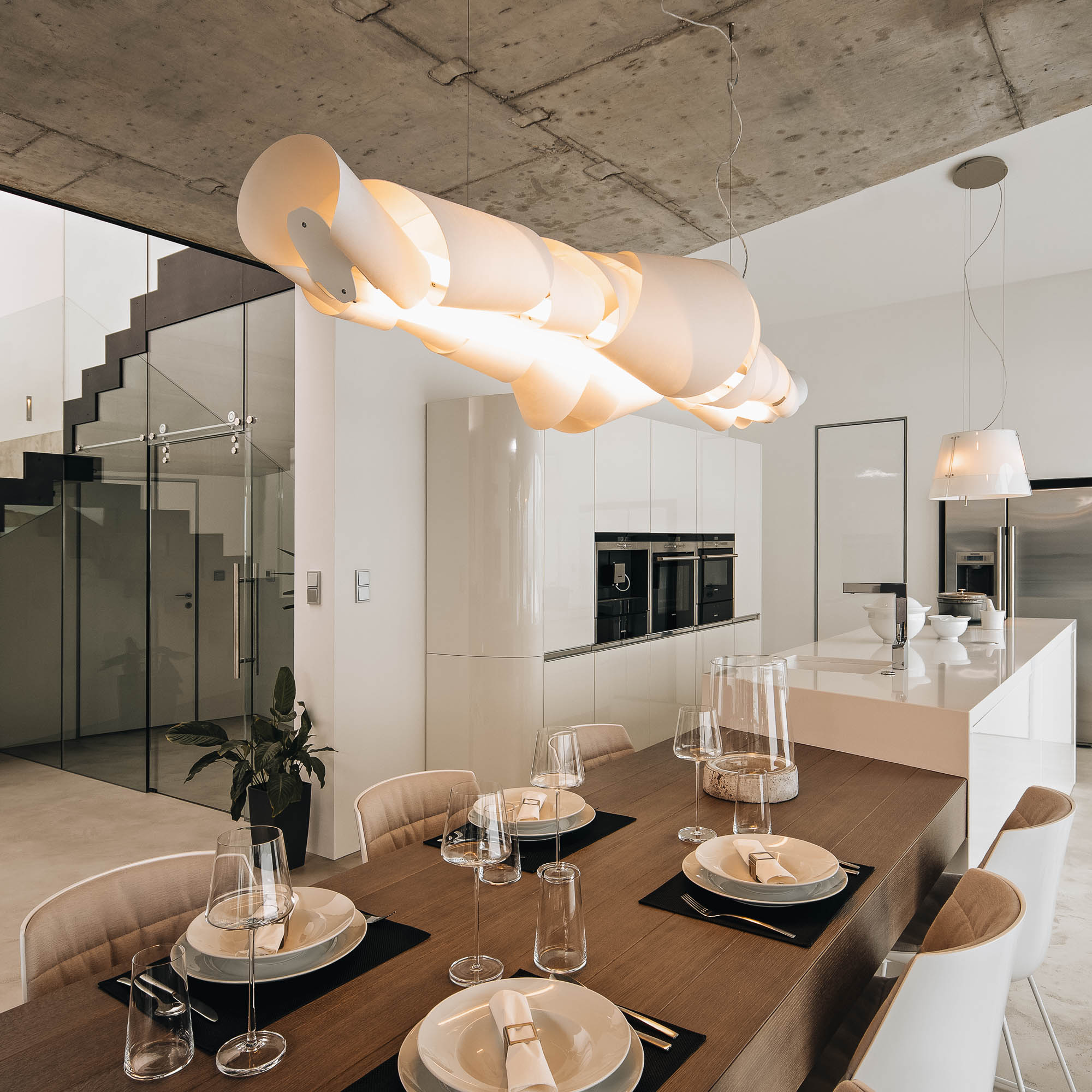 Detail jídelního stolu ve vile s designovým závěsným svítidlem a pohledovým betonovým stropem, OOOOX