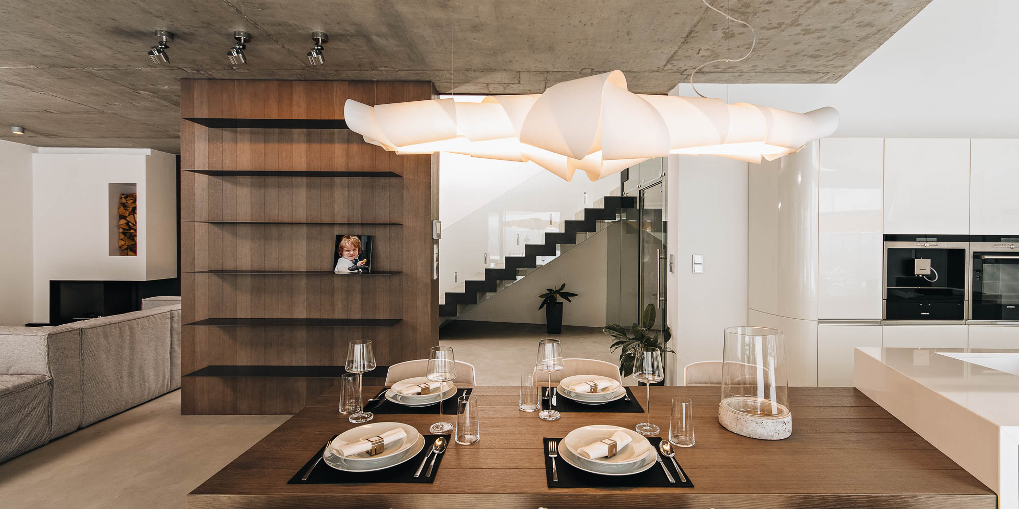 Pohled z kuchyně na jídelnu, obývací pokoj a schodiště v otevřeném prostoru vily, pohledový beton, kuchyně s vykonzolovaným jídelním stolem