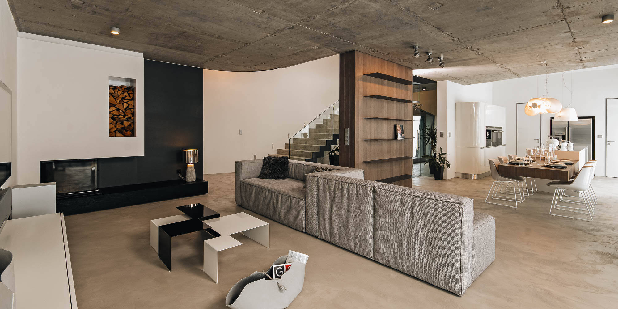 Obývací pokoj vily s krbem, pohledovým betonovým stropem, schodištěm a jídelnou v pozadí, OOOOX