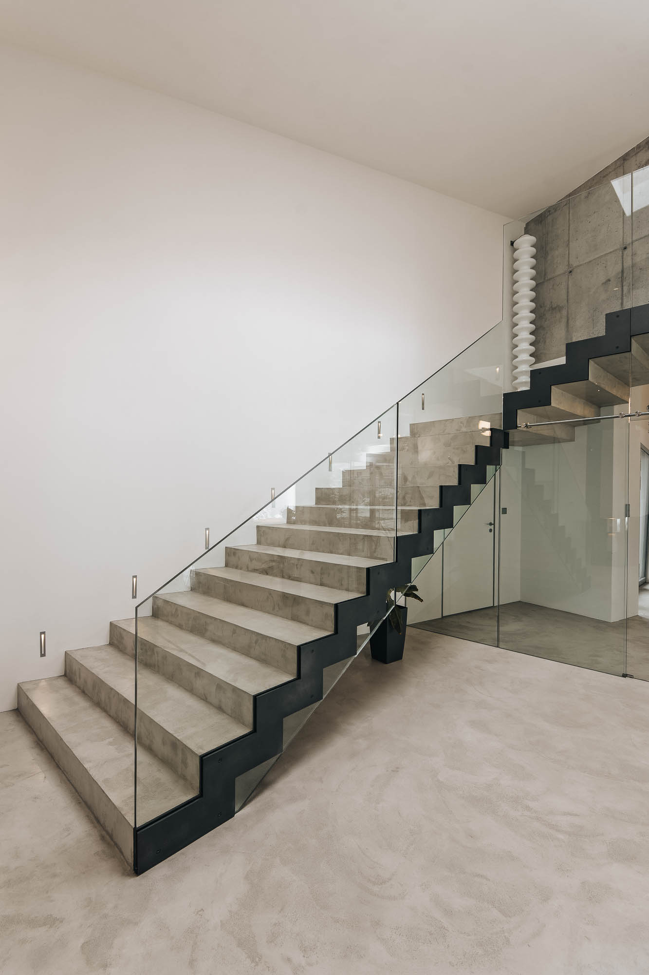 Schodiště vily s pohledovým betonem, skleněným zábradlím a designovým bílým radiátorem na podestě, OOOOX