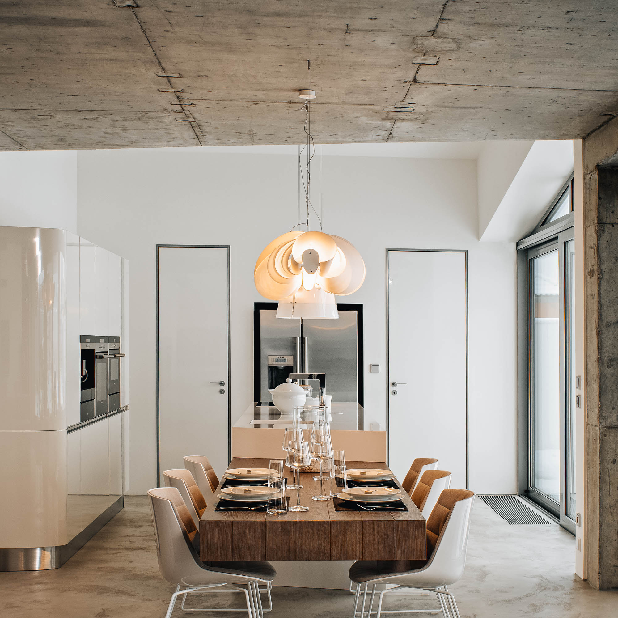 Jídelna vily s bílou lesklou kuchyní, jídelním stolem a designovým závěsným svítidlem, pohledový beton