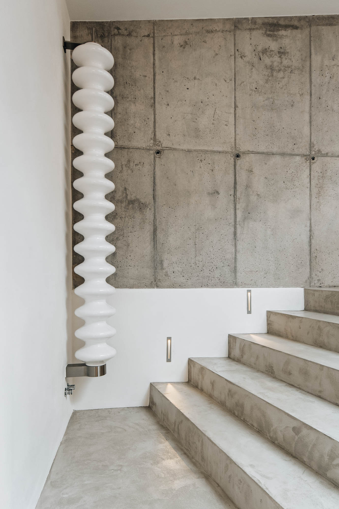 Designový bílý radiátor ve tvaru šroubovice na pohledové betonové stěně u schodiště vily, návrh OOOOX