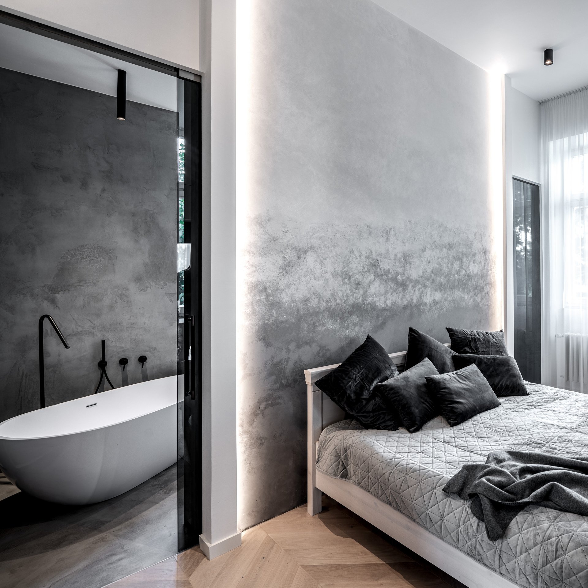 Ložnice s pohledem do en-suite koupelny odkud je vidět volně stojící oválná bílá vana, stěna z benátské omítky s nepřímým světlem. OOOOX