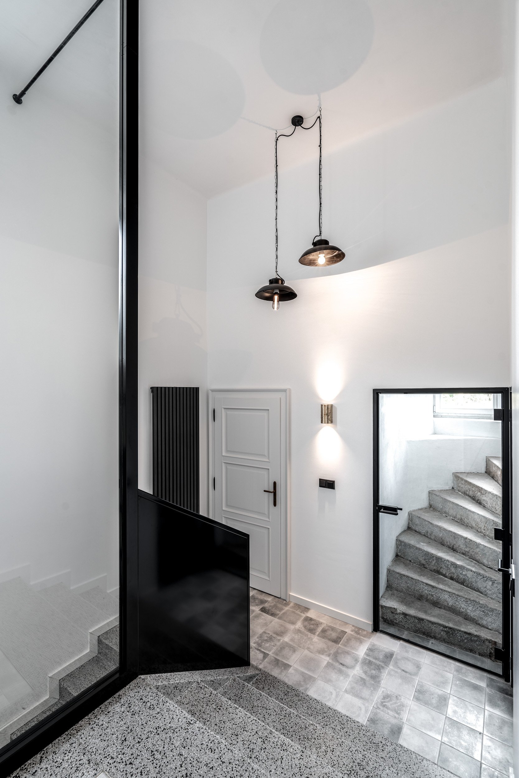 Staircase landing with terrazzo floor, transition to grey stone tiles, industrial pendant lights and black steel-and-glass partition wall. OOOOX