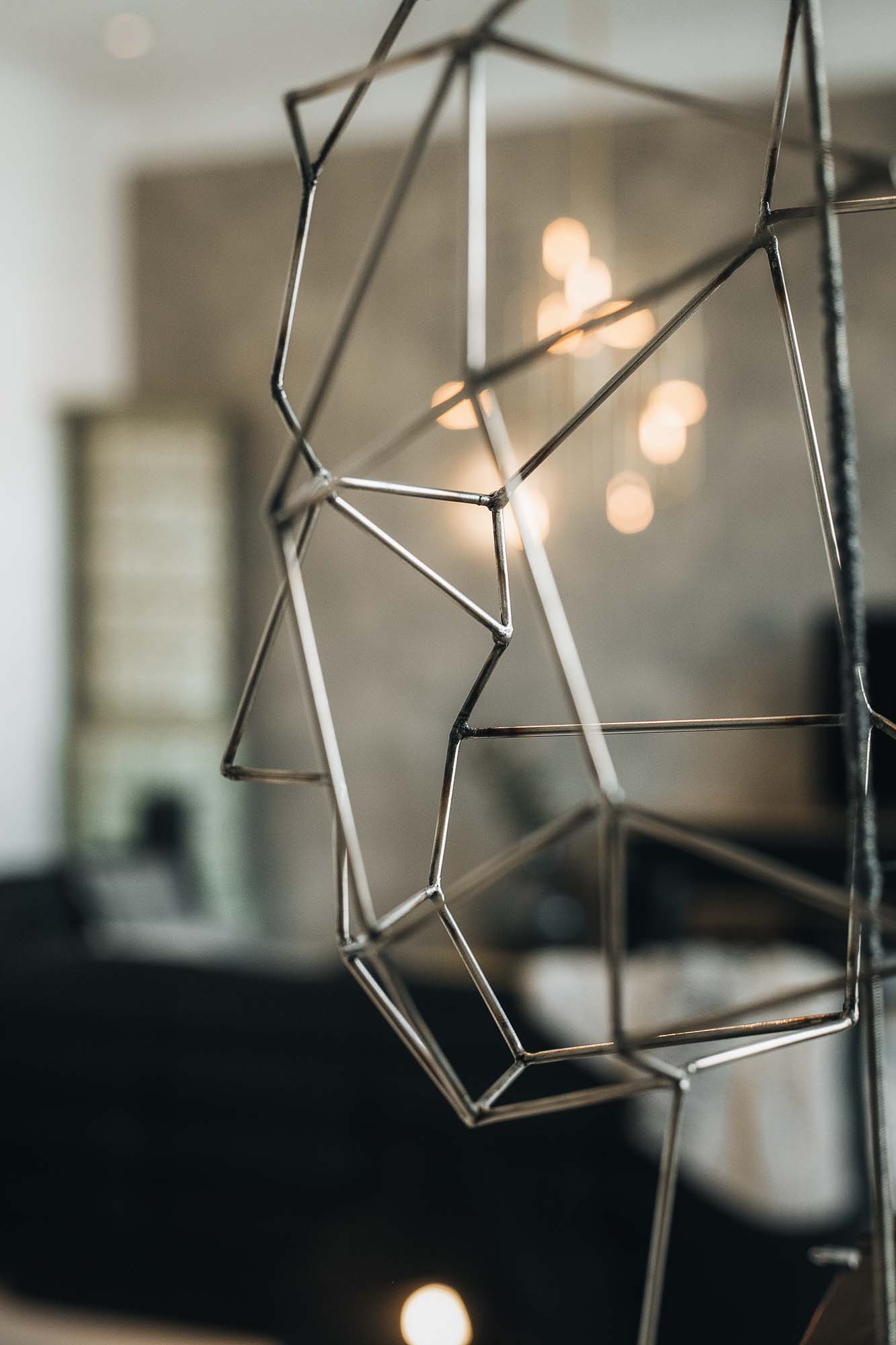 Detailní záběr na geometrický drátěný dekorativní objekt z kovu v bytě Pařížská – abstraktní skulptura z ocelových tyček v popředí, bokeh světel interiéru v pozadí