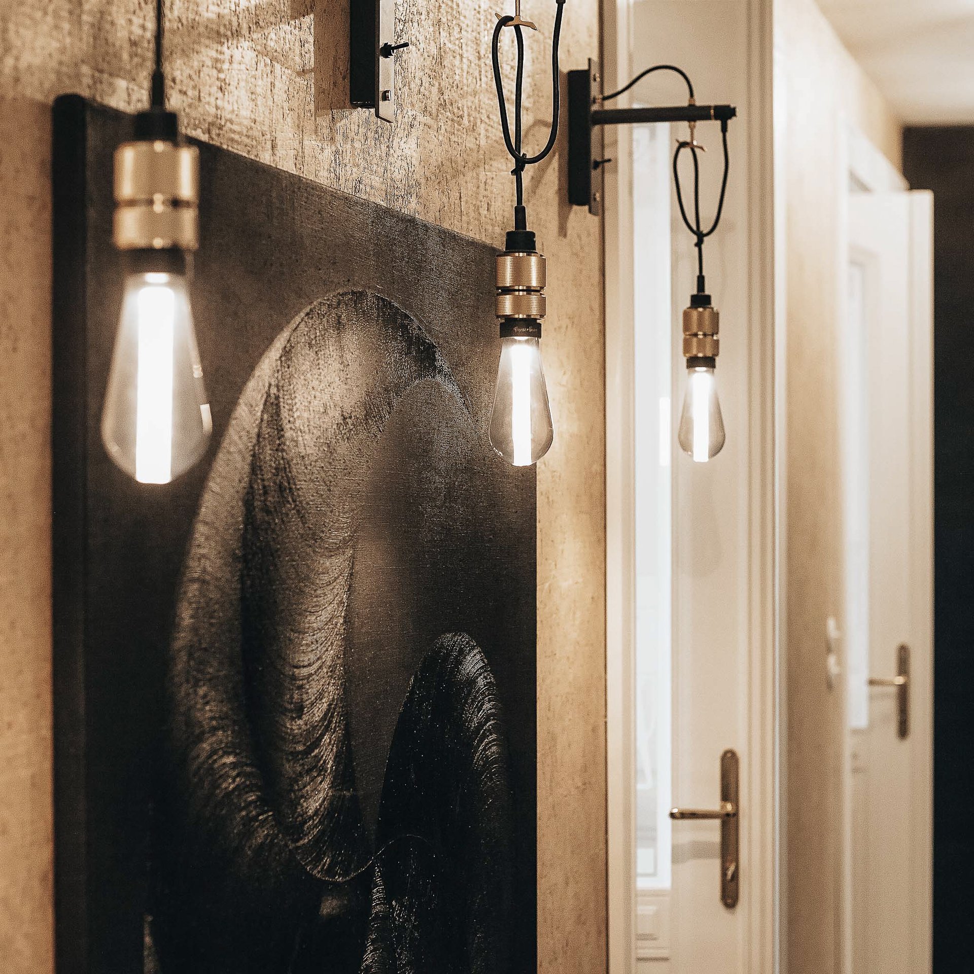 Alternativní záběr chodby bytu Pařížská – tmavý obraz se sloním motivem, mosasné závěsné lampy, průhled do vedlejší místnosti – mírně odlišný střih a teplota barev